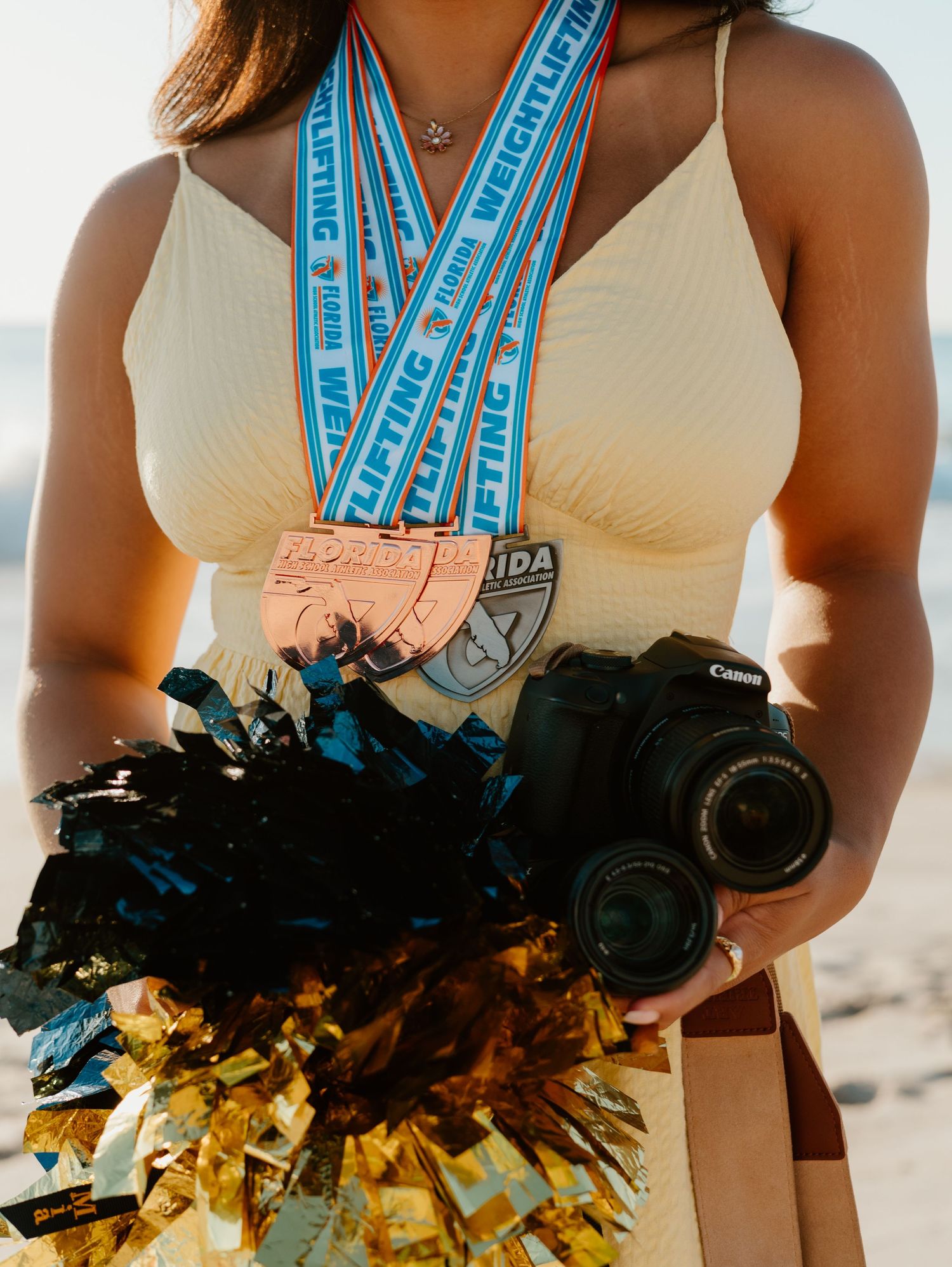 Woman holding Canon camera and pom-poms while wearing weightlifting medals with blue ribbons at a beach.