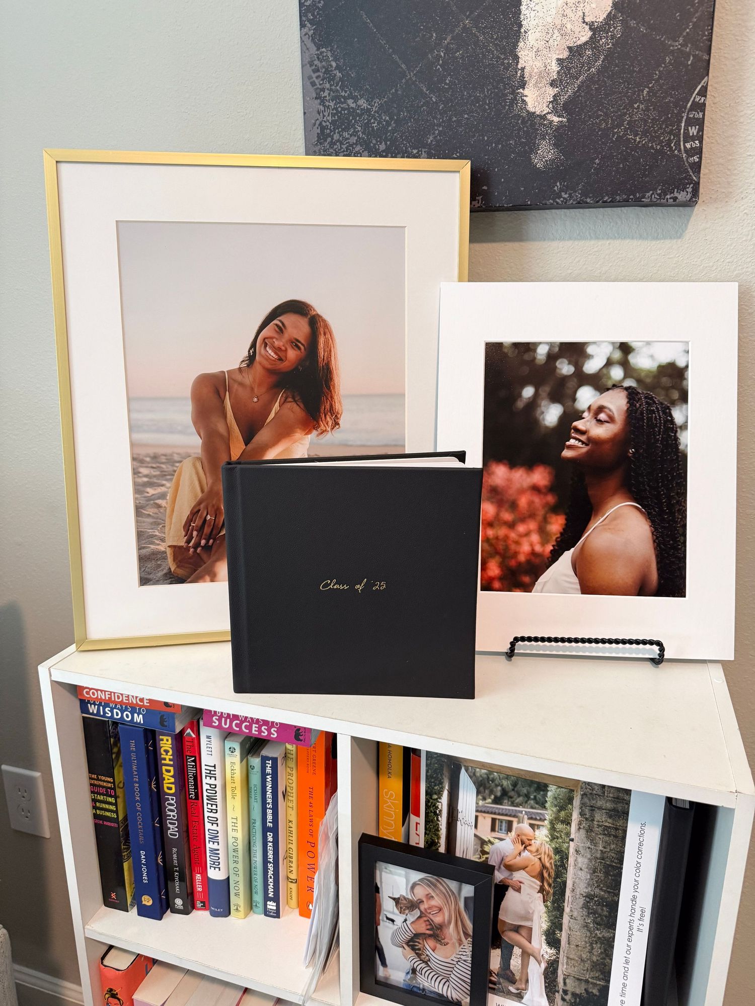 Senior portrait photo album and framed prints displayed on white bookshelf with colorful books, featuring Class of 23 graduation photos.