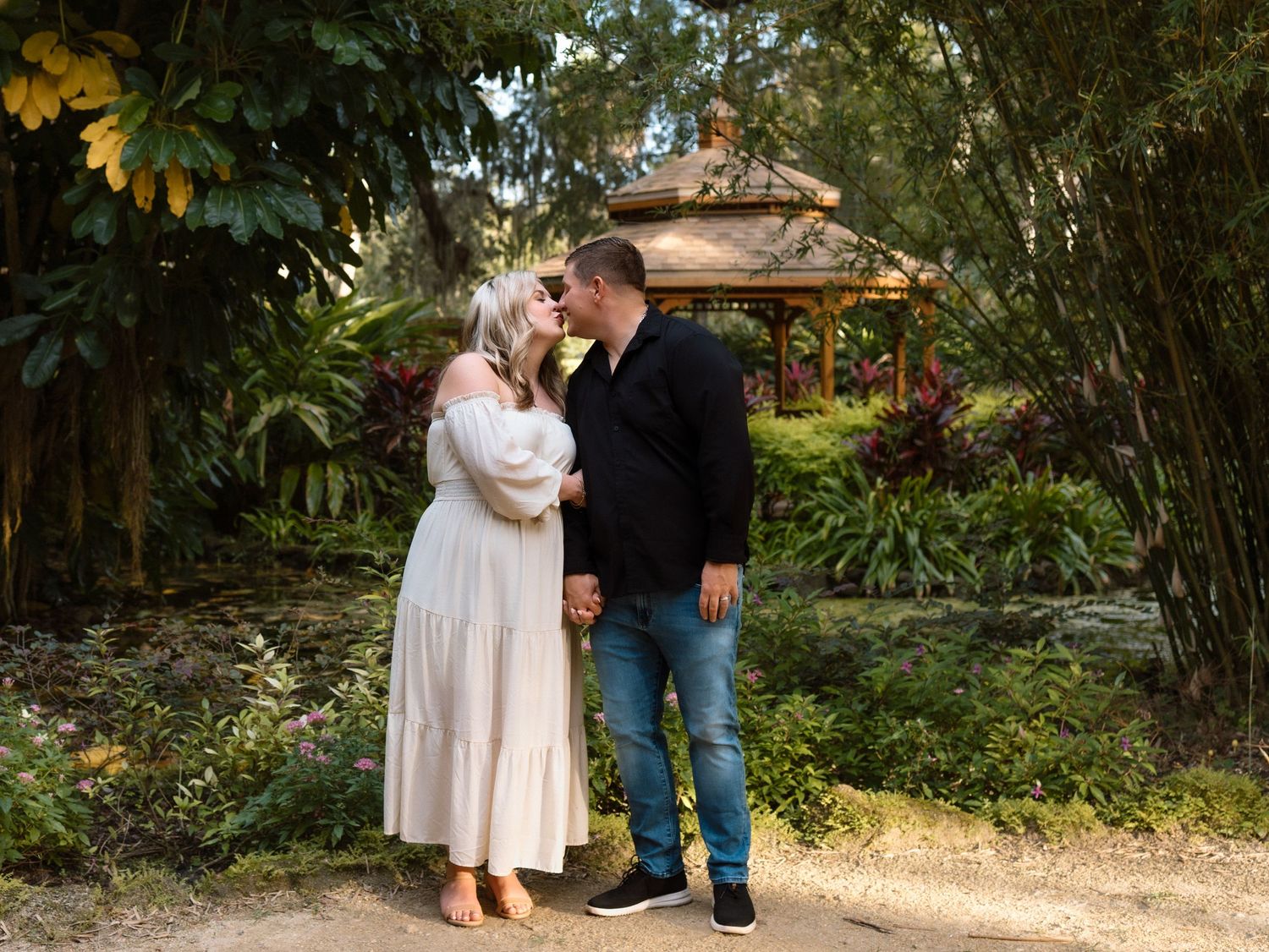Couple sharing a romantic kiss in a lush garden with a gazebo backdrop, woman in white dress, man in black shirt.