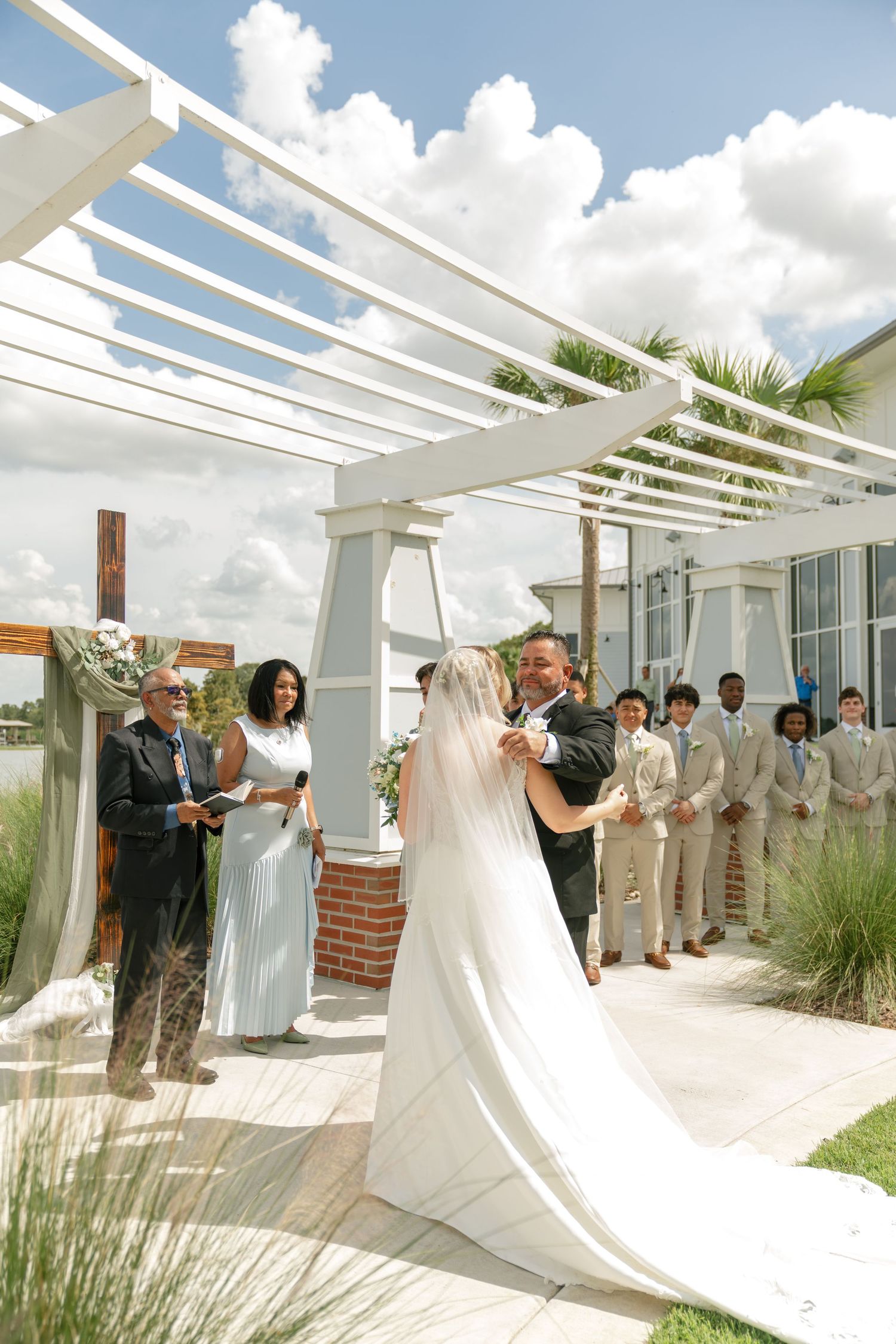 Outdoor wedding ceremony under white pergola with bride, groom, groomsmen in tan suits, and officiant on sunny day.