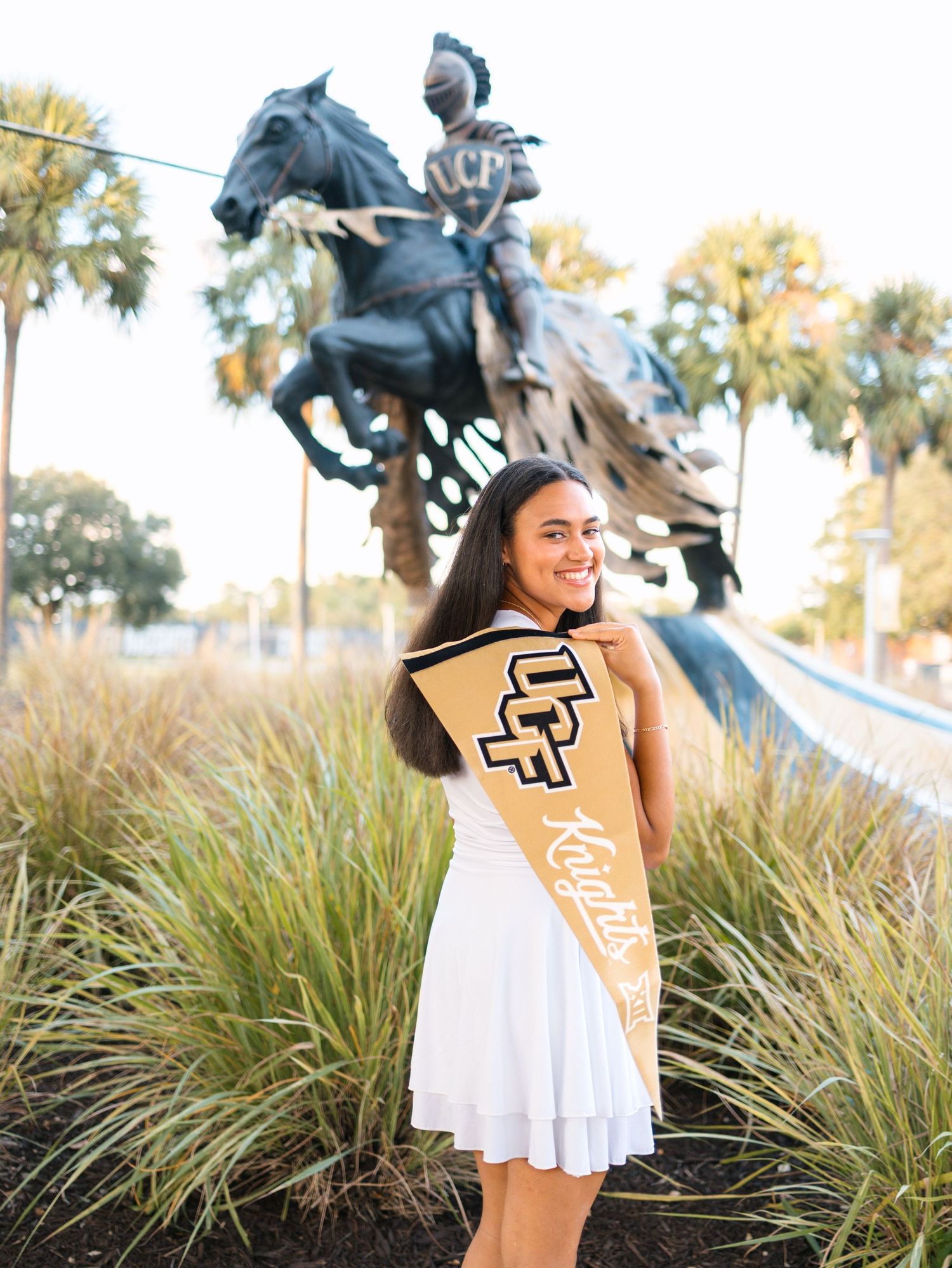 A smiling young woman in a white dress holds a UCF pennant and sash in front of the UCF knight statue on campus.