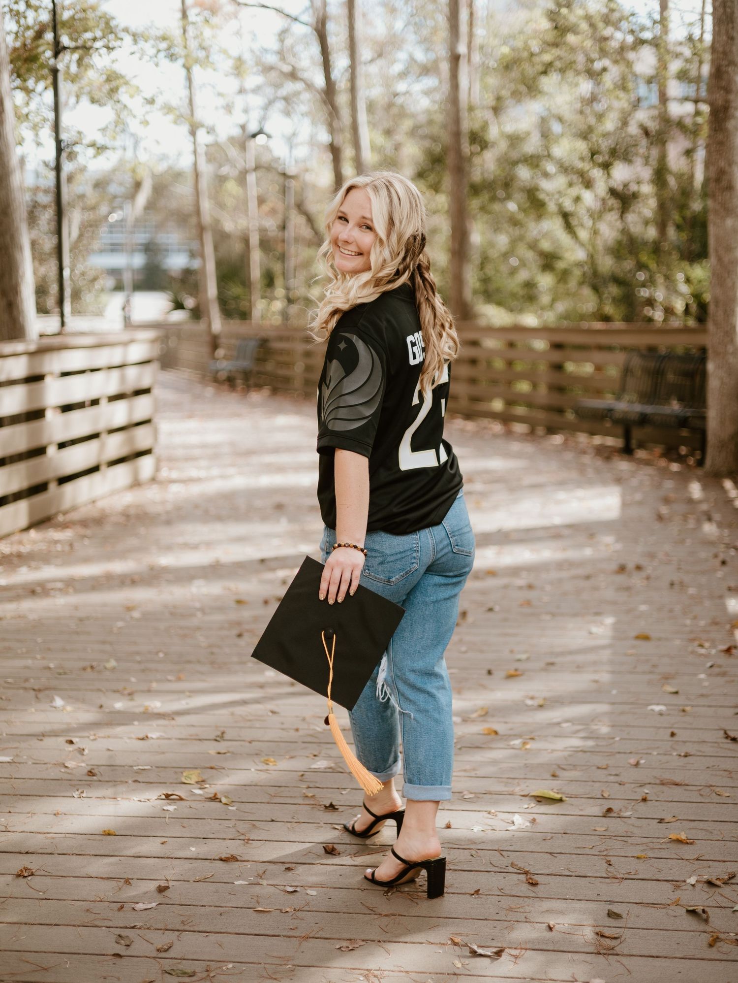 Smiling blonde graduate holding mortarboard cap in jersey and jeans on wooden bridge outdoors.