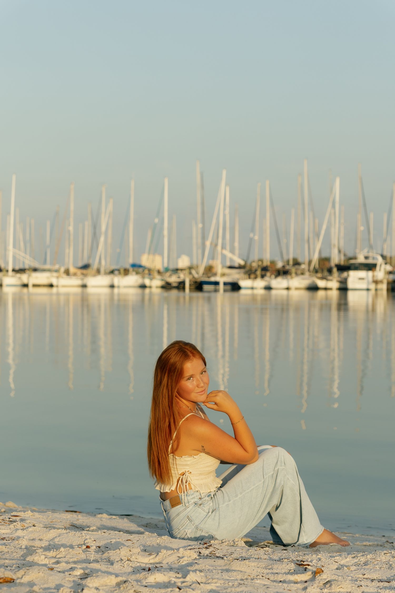 Young redhead woman sitting barefoot on sandy beach shore with marina sailboats reflecting in calm water at sunset.