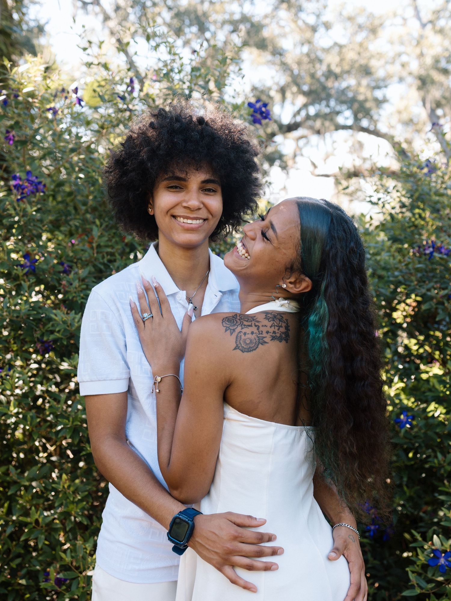 A couple wearing white outfits embraces and laughs together in a garden setting with flowering bushes in the background.