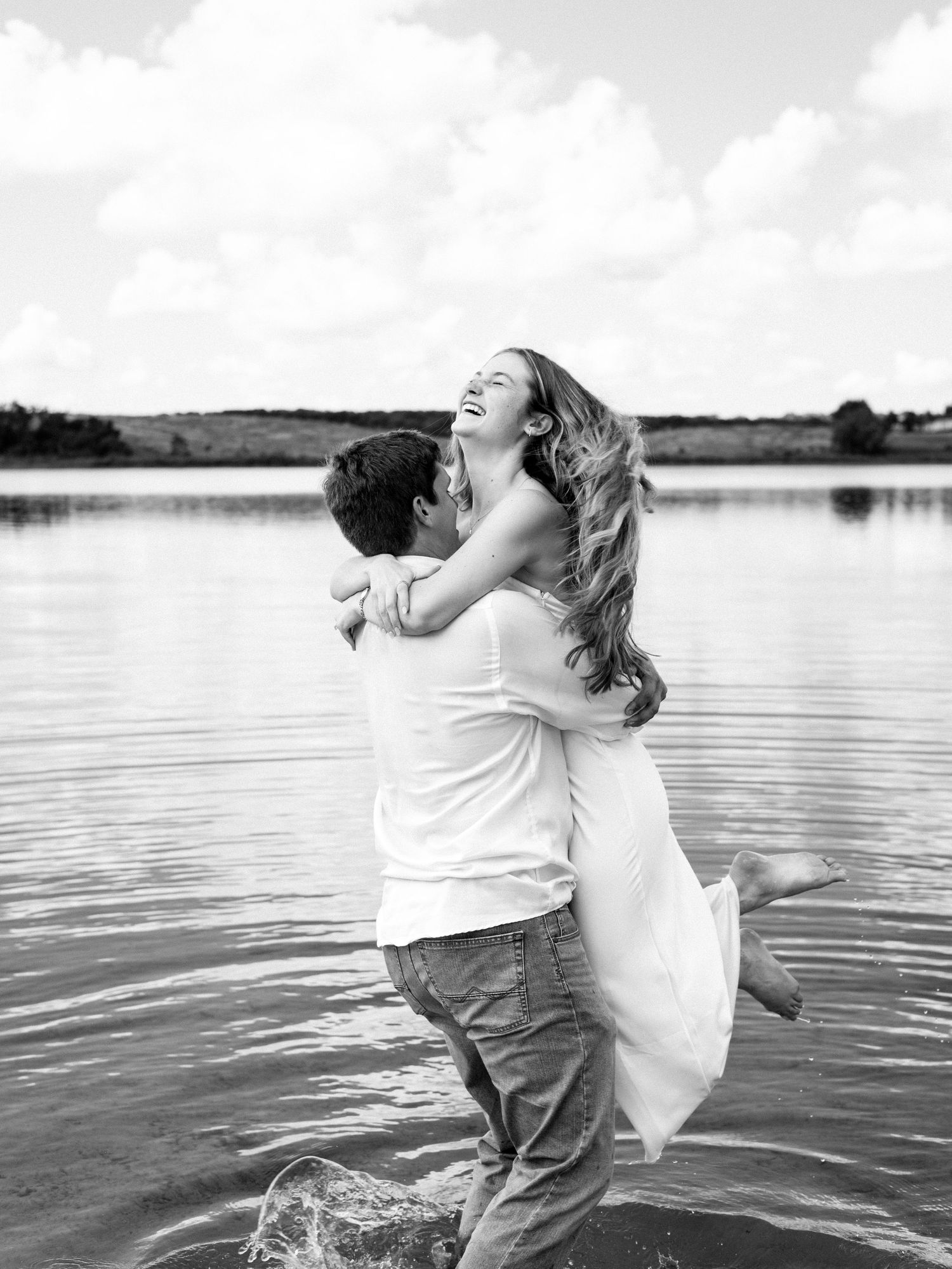 A couple dances playfully together in shallow lake water, creating a romantic black and white silhouette against the horizon.