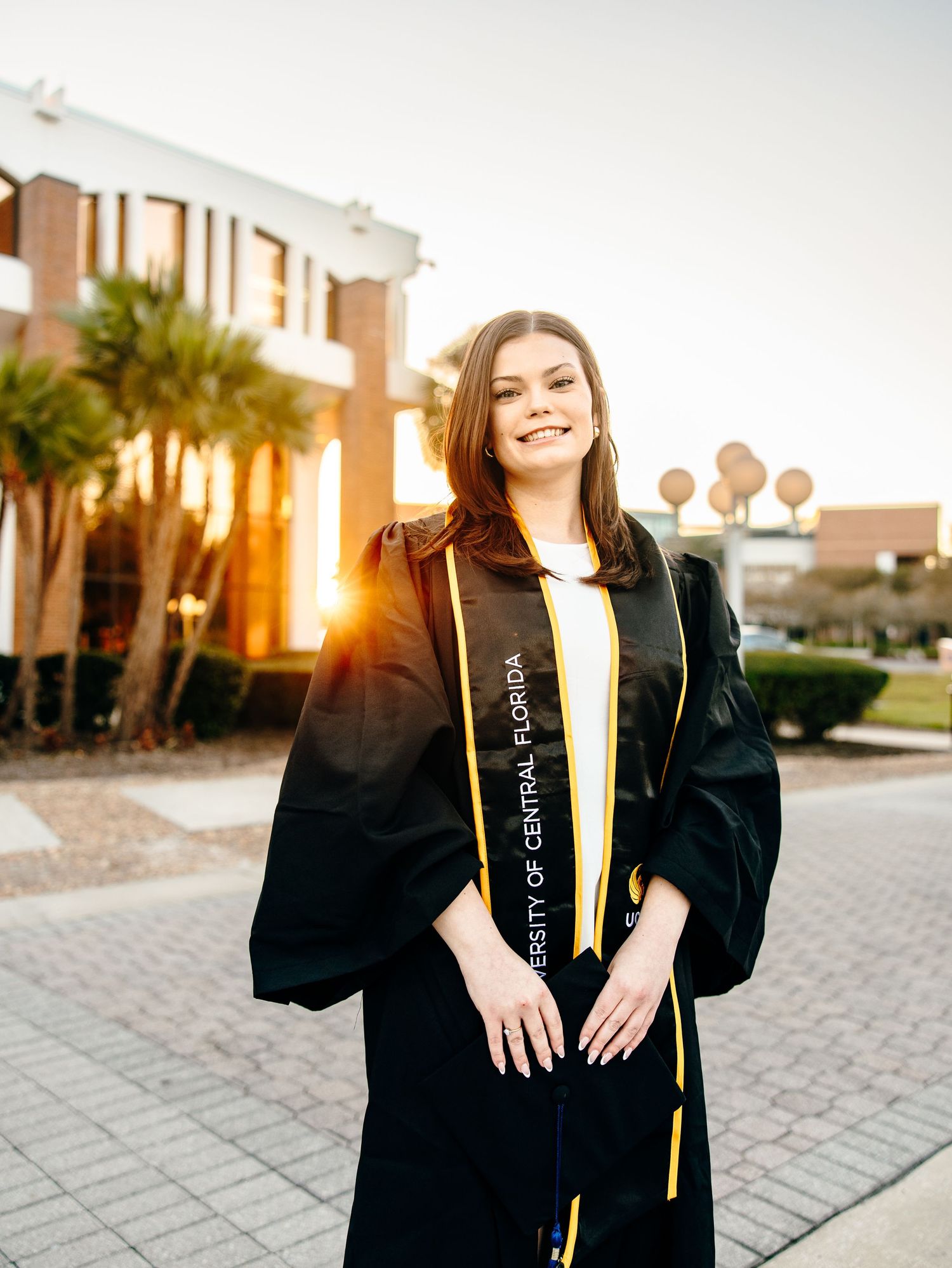 UCF graduate in black cap and gown with gold sash smiles outdoors on campus at golden hour.