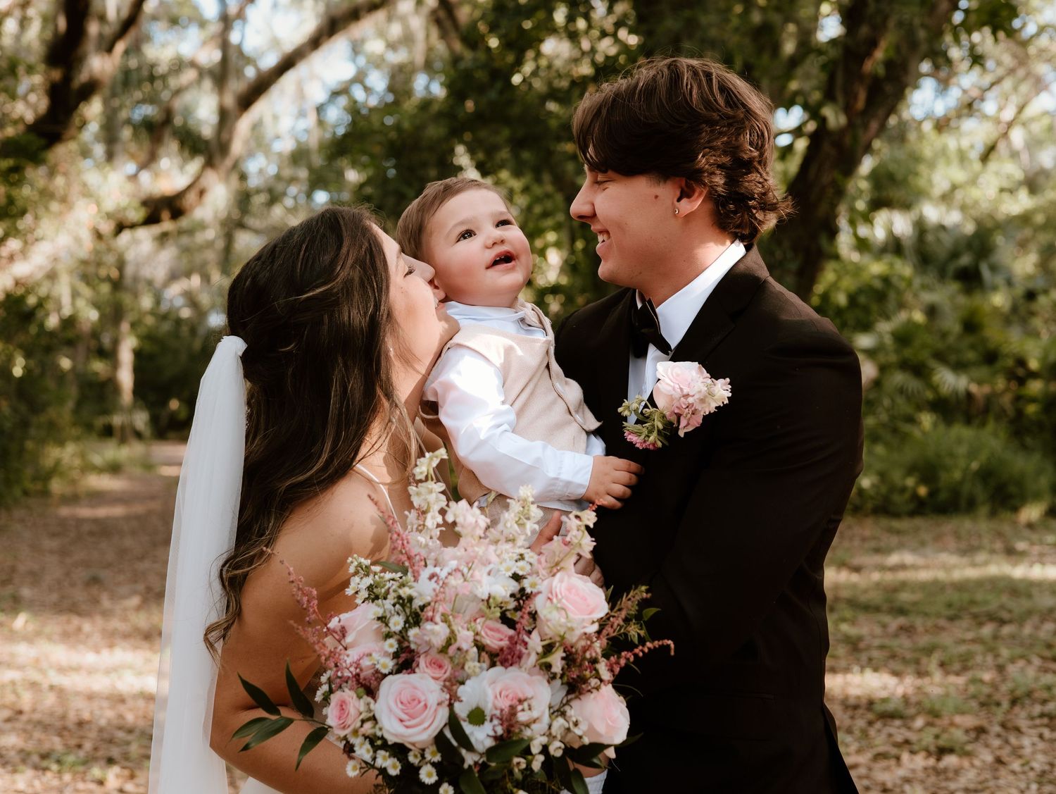 A happy couple in wedding attire shares tender moments while holding their baby in an outdoor setting with trees.