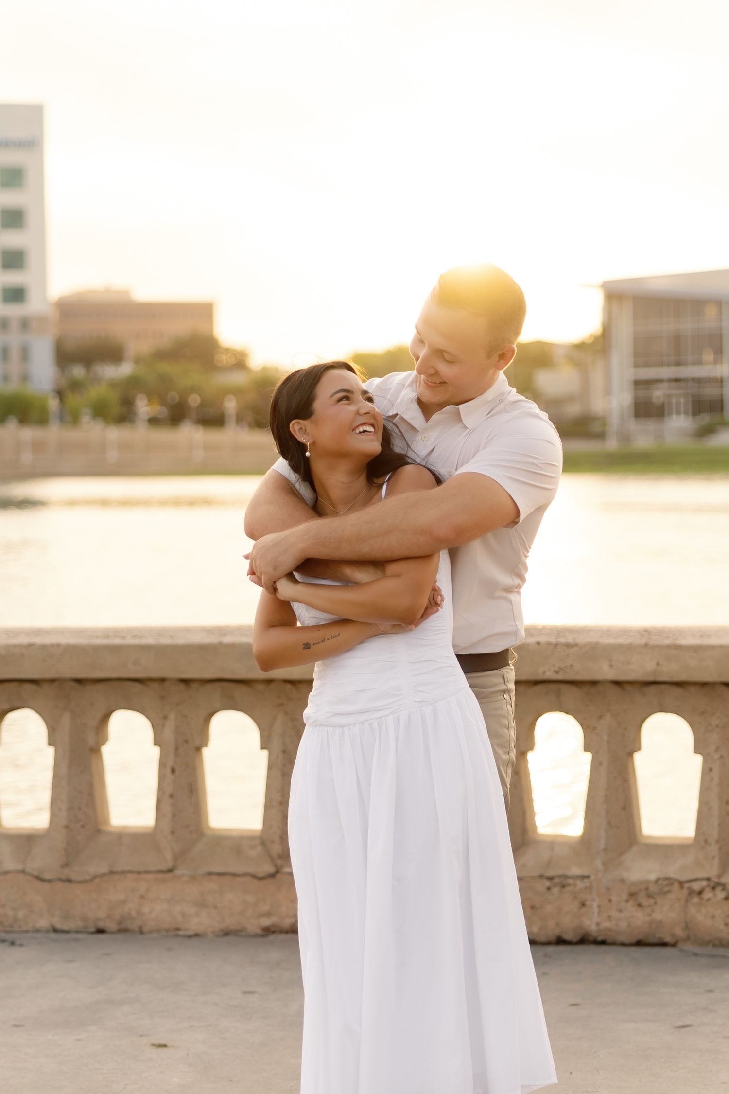 Couple embracing at sunset by a waterfront balustrade, woman in white dress smiling up at man in warm golden light.