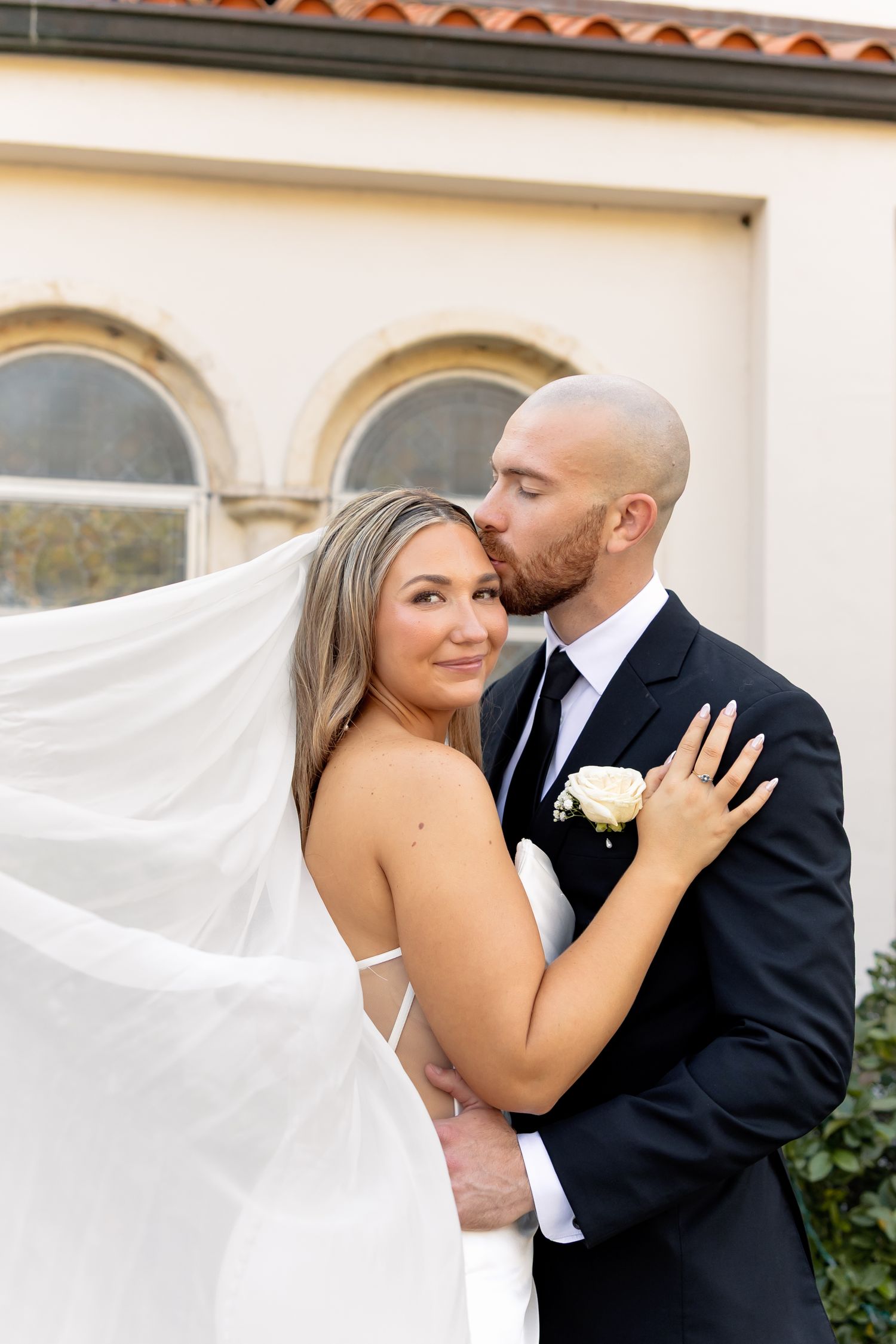 Groom in black tuxedo kissing bride on forehead while embracing her flowing white veil outdoors.