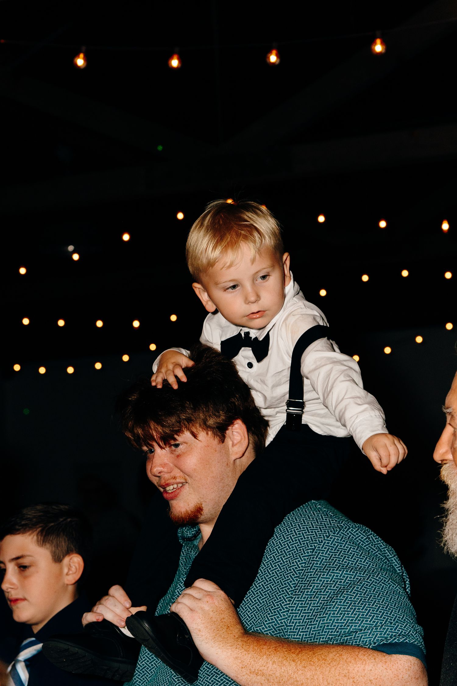 Young boy in suspenders and bow tie sitting on mans shoulders at a wedding reception.
