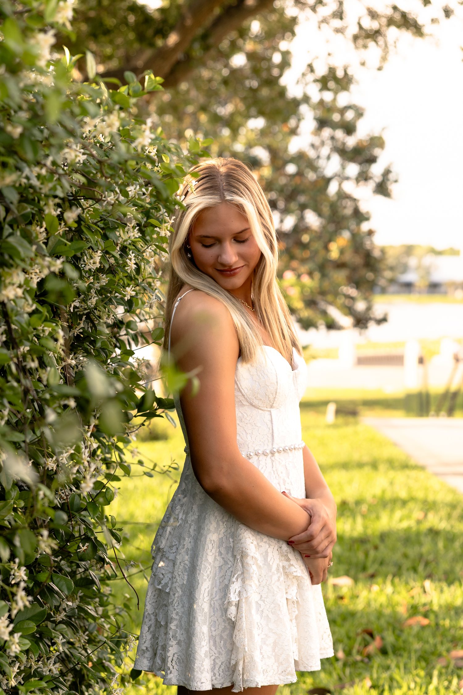Young blonde woman in white lace dress smiling outdoors near flowering bushes in warm sunlight.