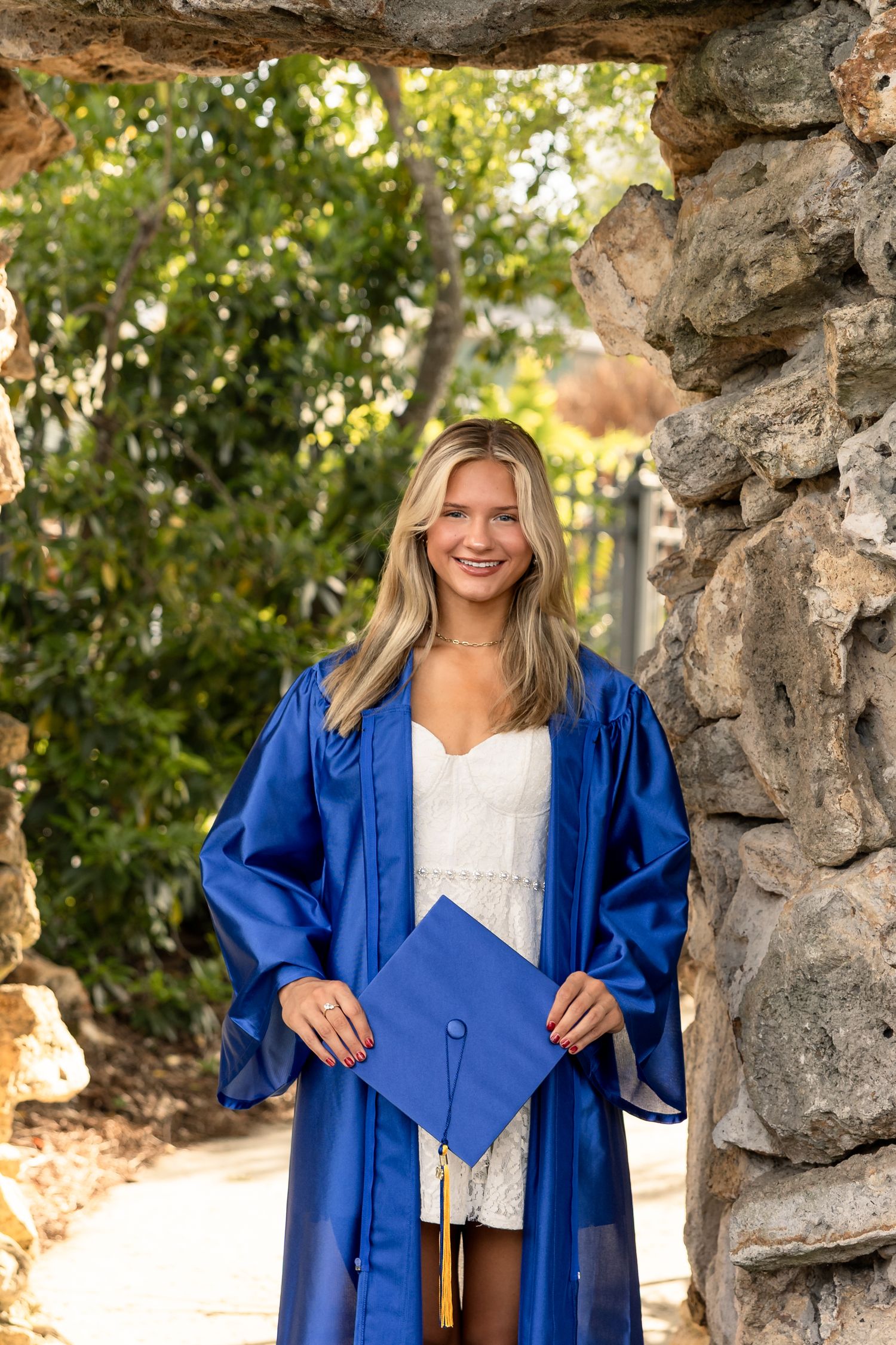 Smiling blonde graduate in blue cap and gown poses outdoors near stone wall with lush greenery backdrop.