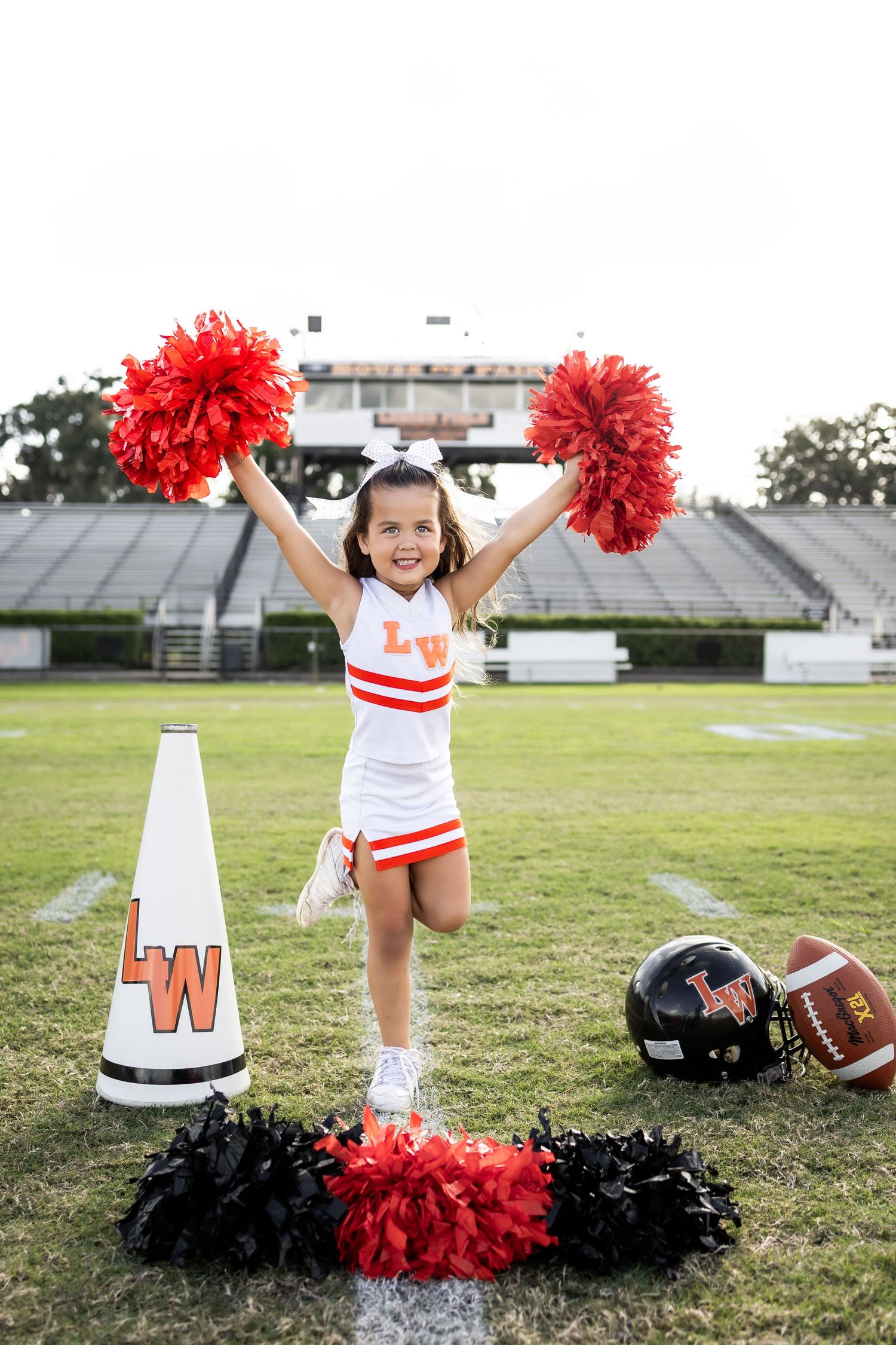 Young girl in LW cheerleader uniform holds red pom-poms on football field with megaphone and helmet nearby.