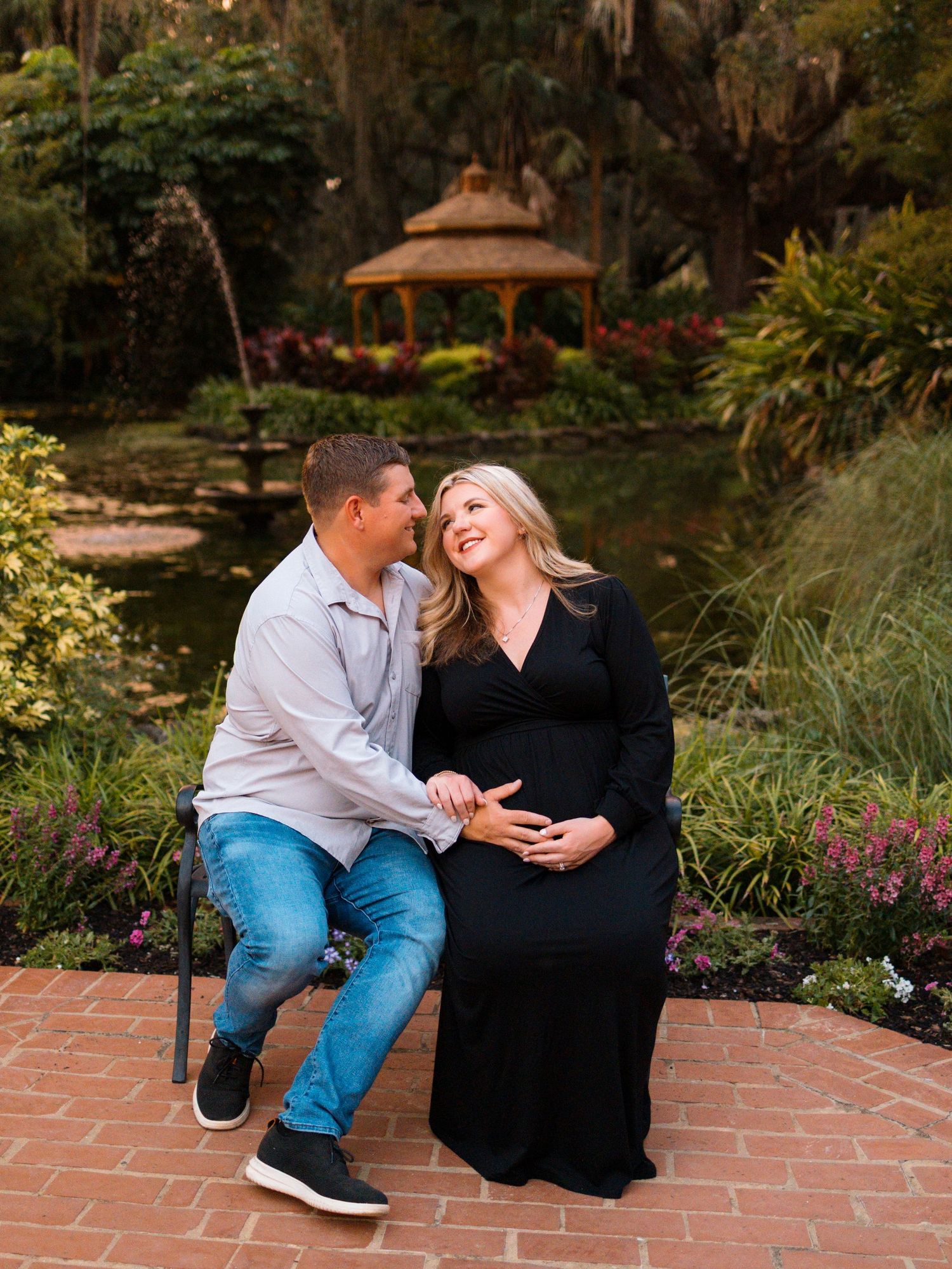 Expecting couple sitting together in a lush garden near a gazebo, woman in black maternity dress smiling joyfully.