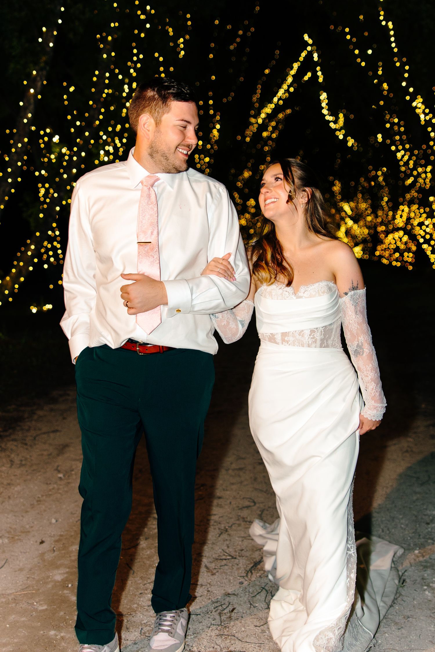 Bride in white lace gown and groom in white shirt with pink tie walk together under golden fairy lights at night.