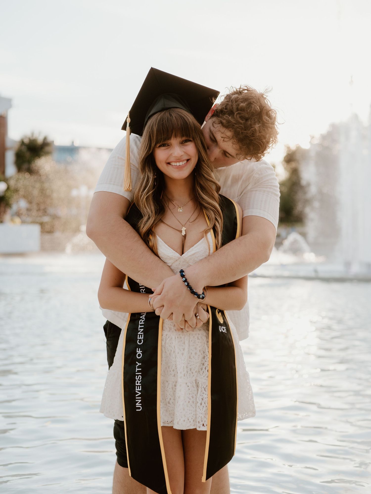A smiling graduate in cap and stole from University of Central Florida embraces her partner near a fountain.