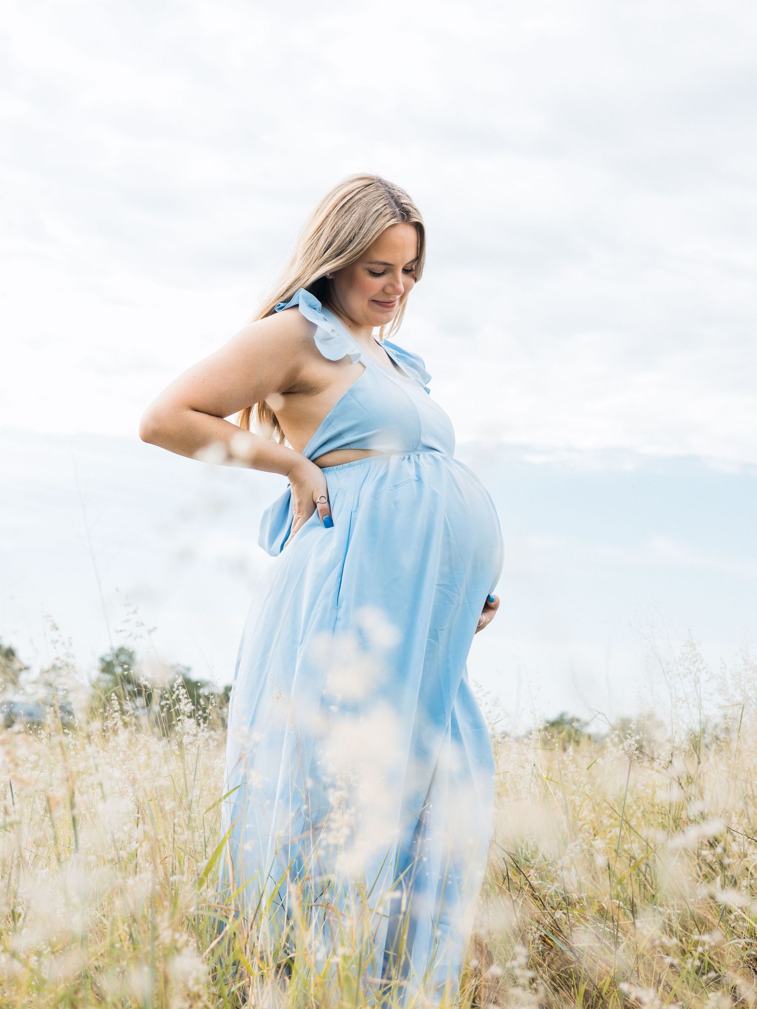 Someone in a flowing light blue dress stands in a sunlit meadow of tall grass against a cloudy sky.
