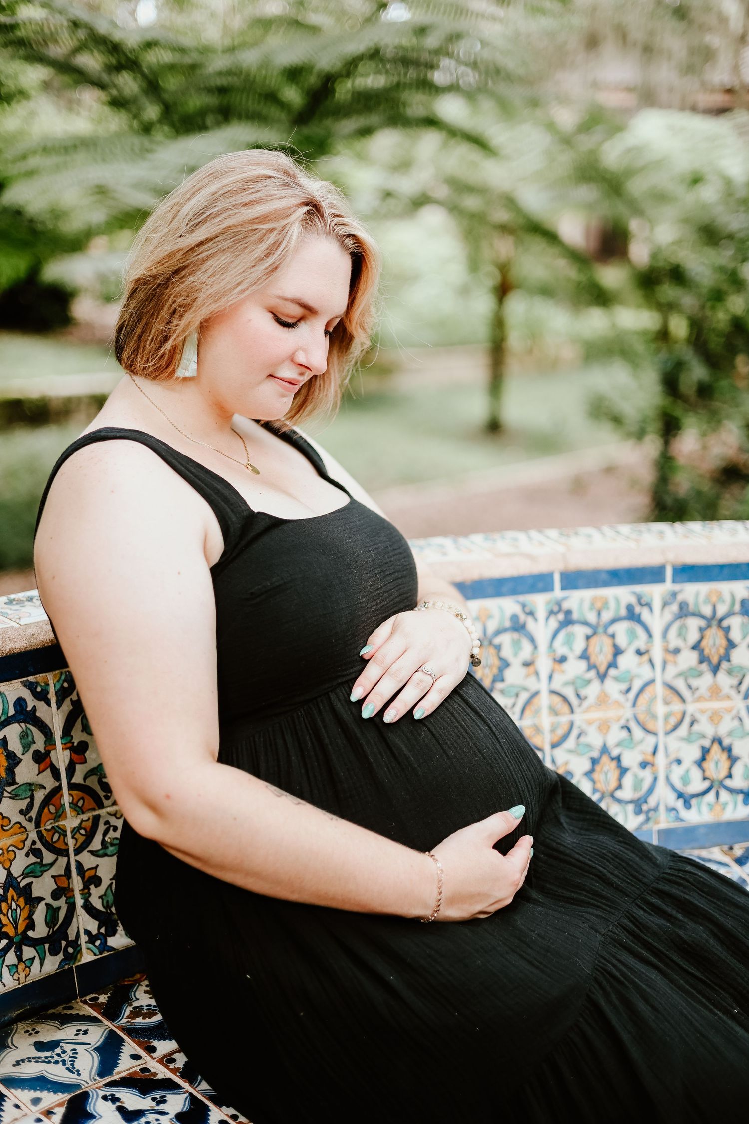 A pregnant woman in a flowing black dress sits on a decorative blue and white tiled bench in a garden setting.