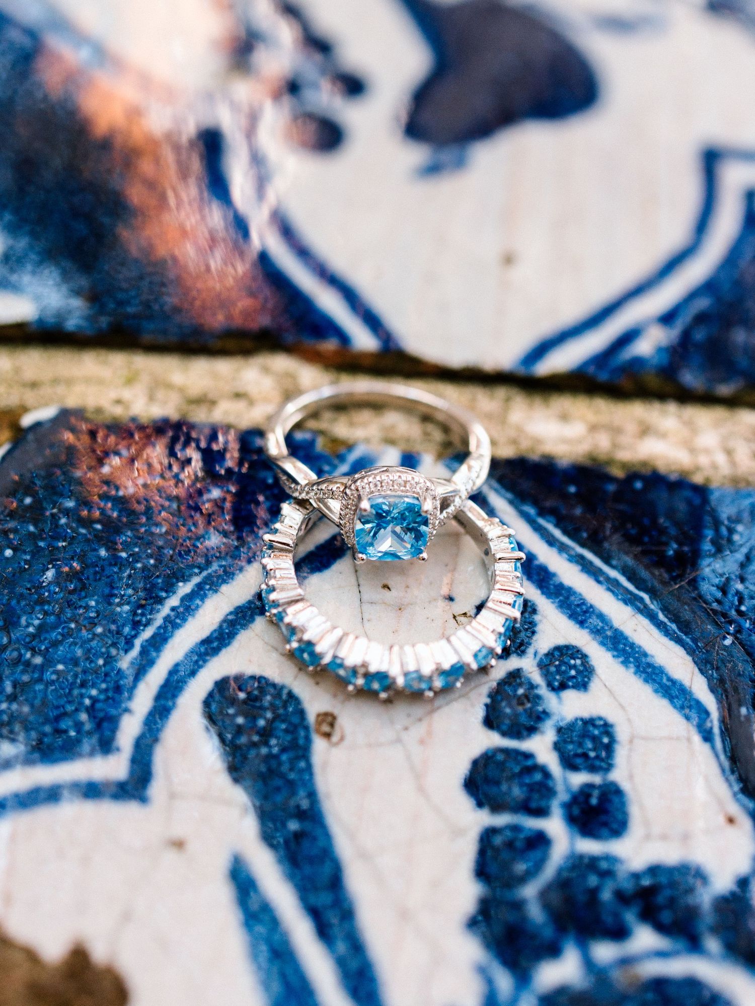 Elegant engagement ring with blue topaz stone and diamond halo displayed on blue and white patterned background.