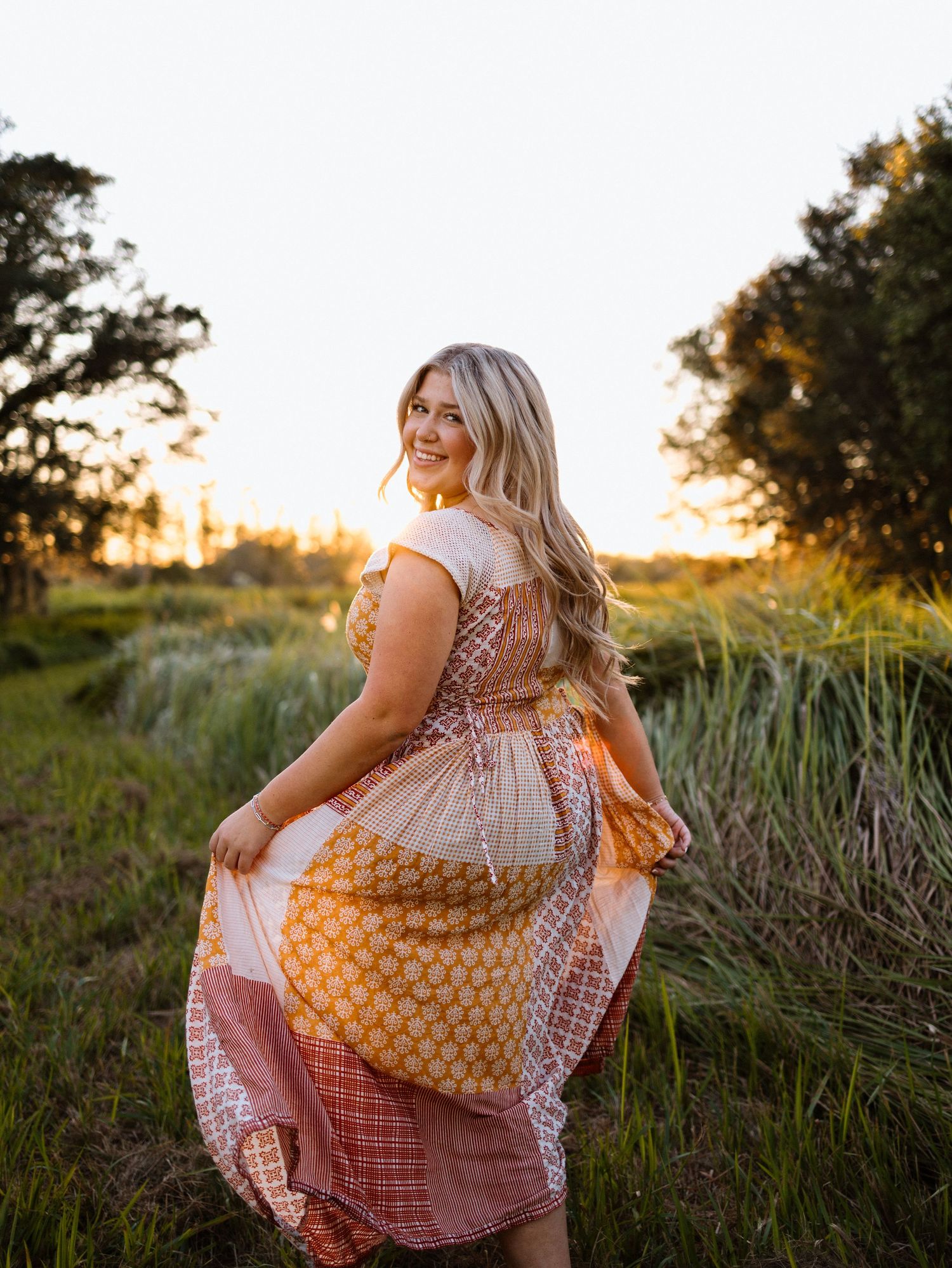 A person in a flowy dress sits in a field of tall grass and wildflowers during golden hour sunset.