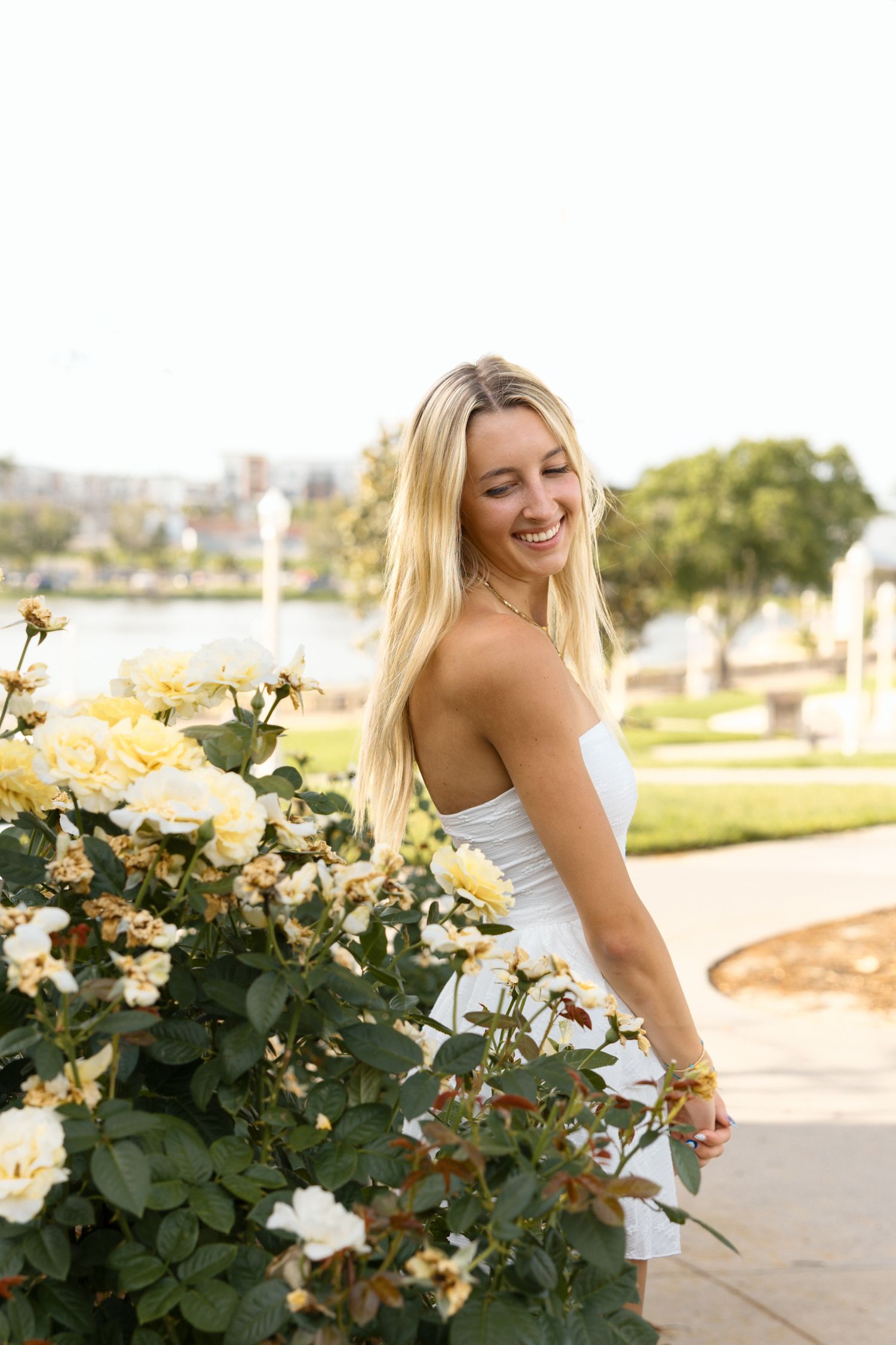 Smiling blonde woman in white dress posing beside yellow roses in a sunny outdoor park setting.