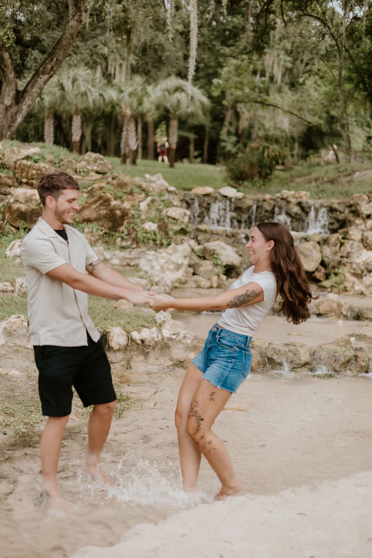 A smiling couple holds hands and splashes in shallow water near a rocky waterfall surrounded by lush tropical greenery.