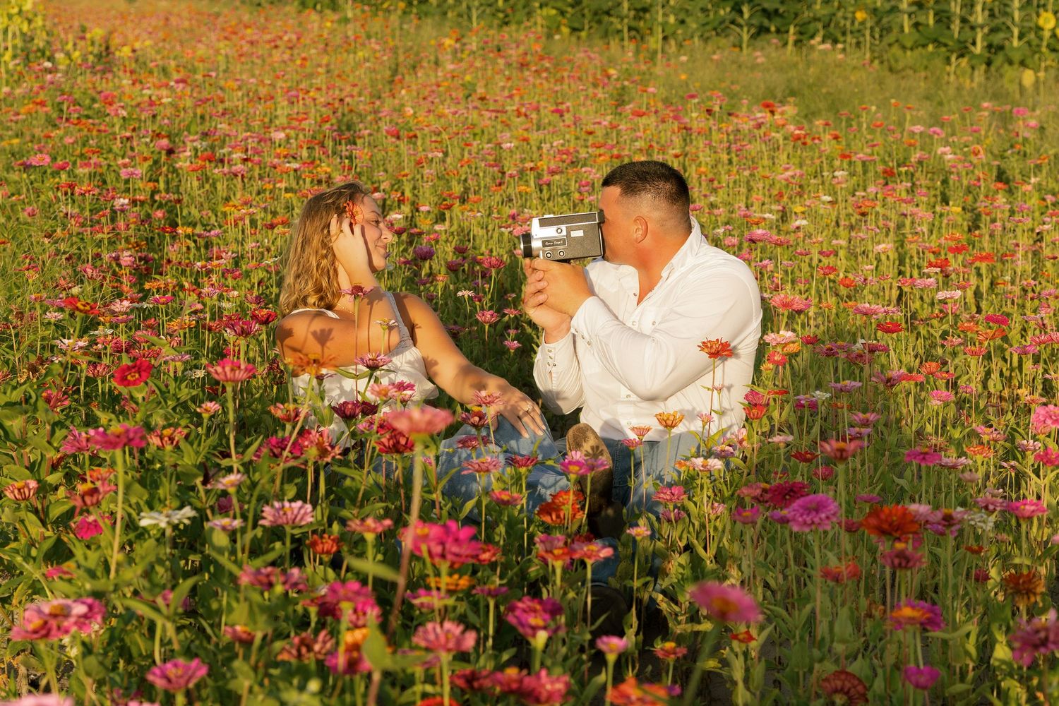 Couple sitting in a vibrant flower field, man filming woman with a video camera among pink and red blooms in golden light.
