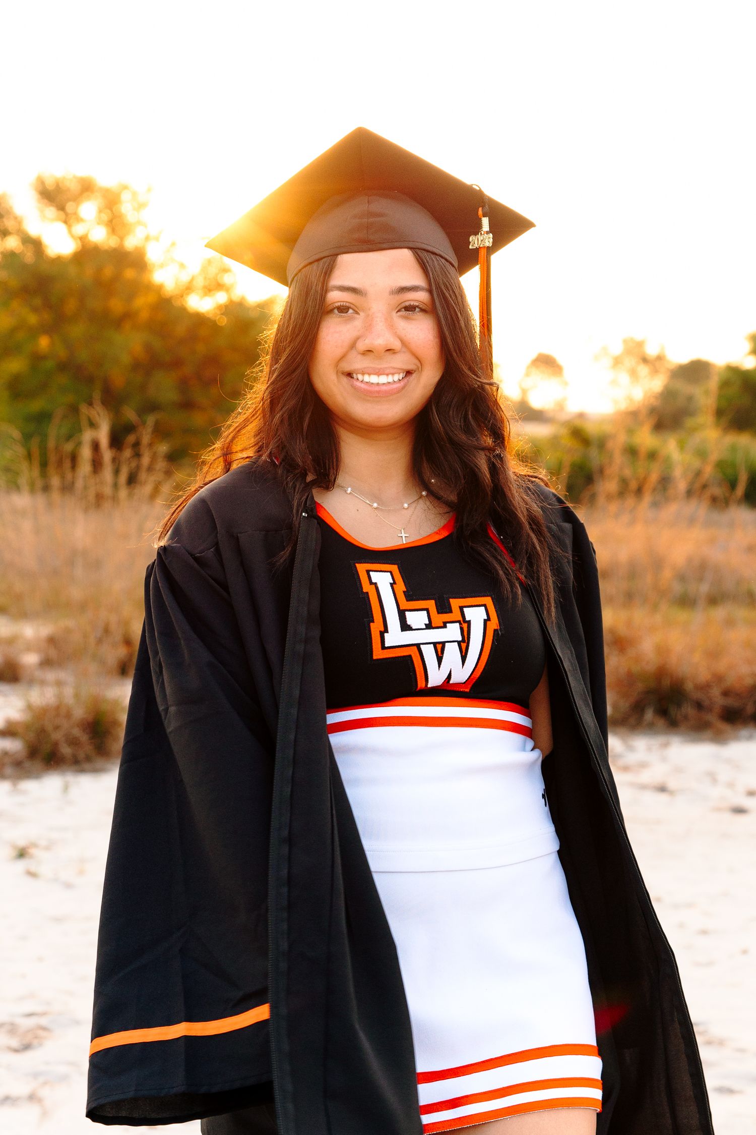 Smiling graduate in black cap and gown over LW cheerleader uniform poses outdoors in golden sunset light.