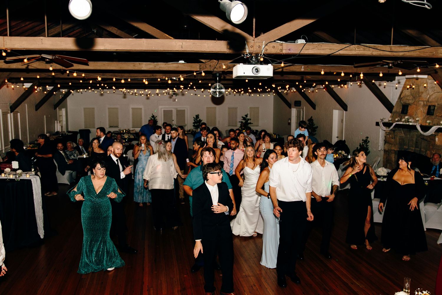 Guests dancing at an elegant wedding reception in a decorated venue with string lights and a wooden ceiling.