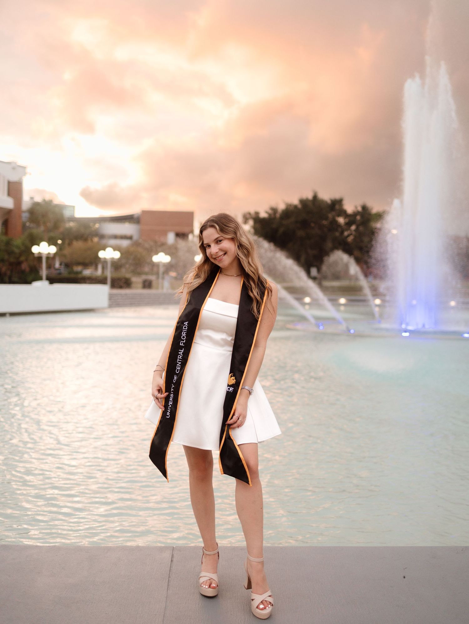 Young woman in white dress and graduation stole poses by illuminated fountain at sunset on university campus.