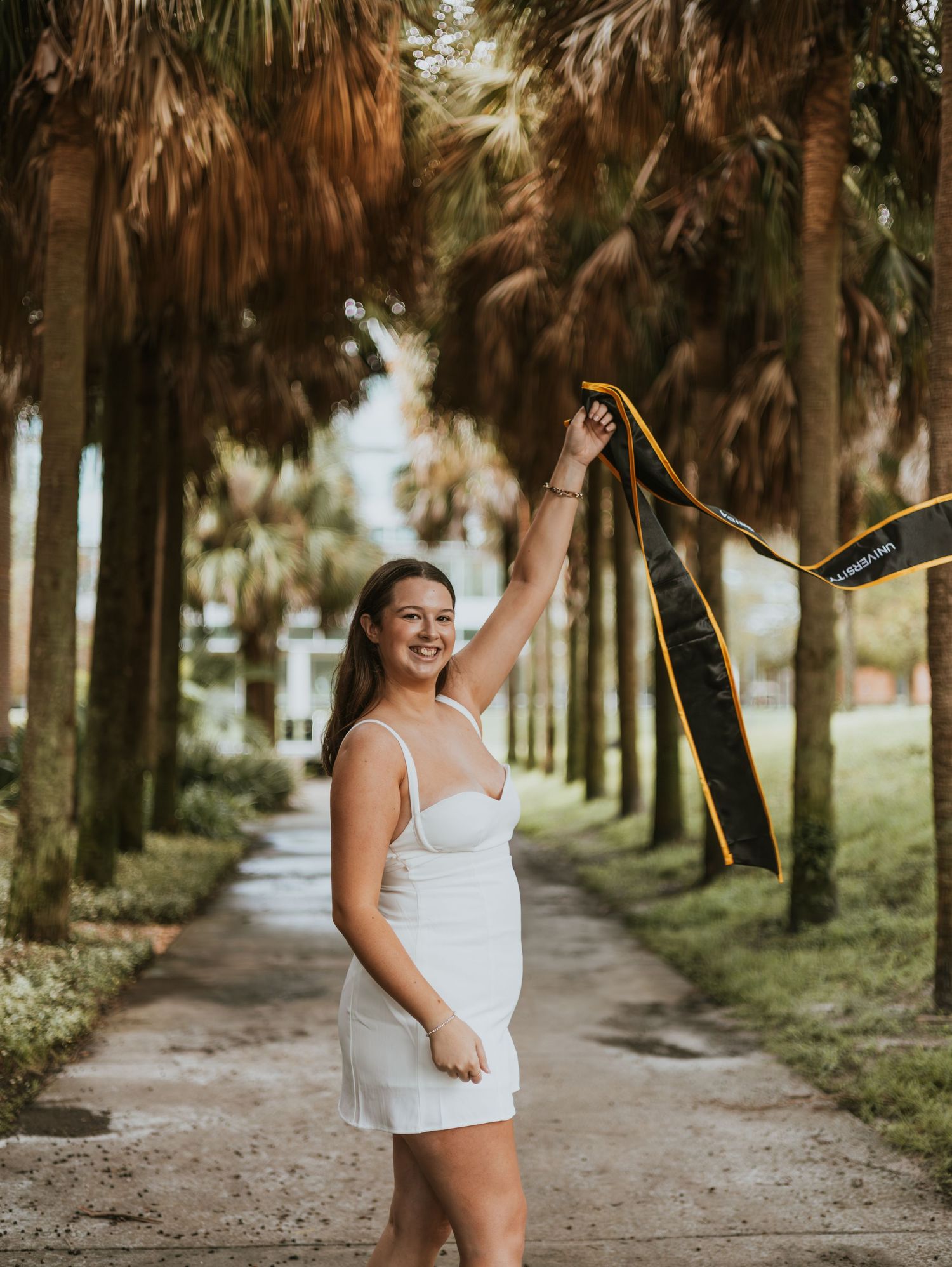 Young woman in white dress smiling and holding a black and gold ribbon aloft on a palm tree-lined path.