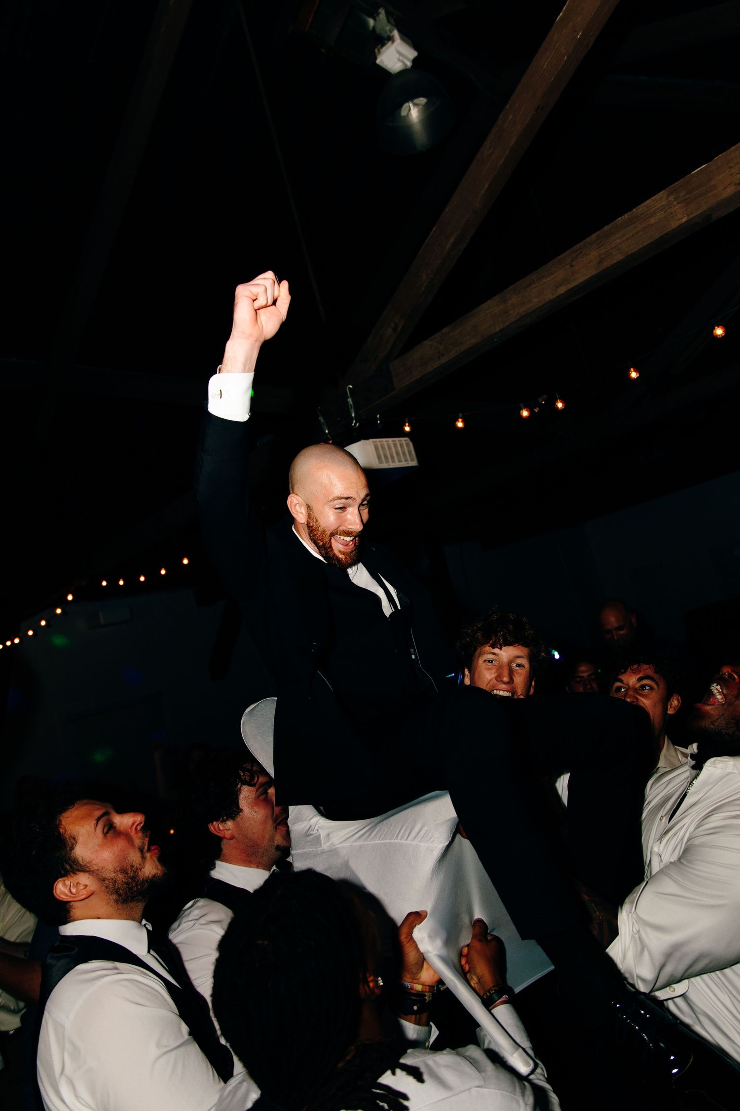 Groom in black tuxedo raised by groomsmen celebrating at wedding reception party.