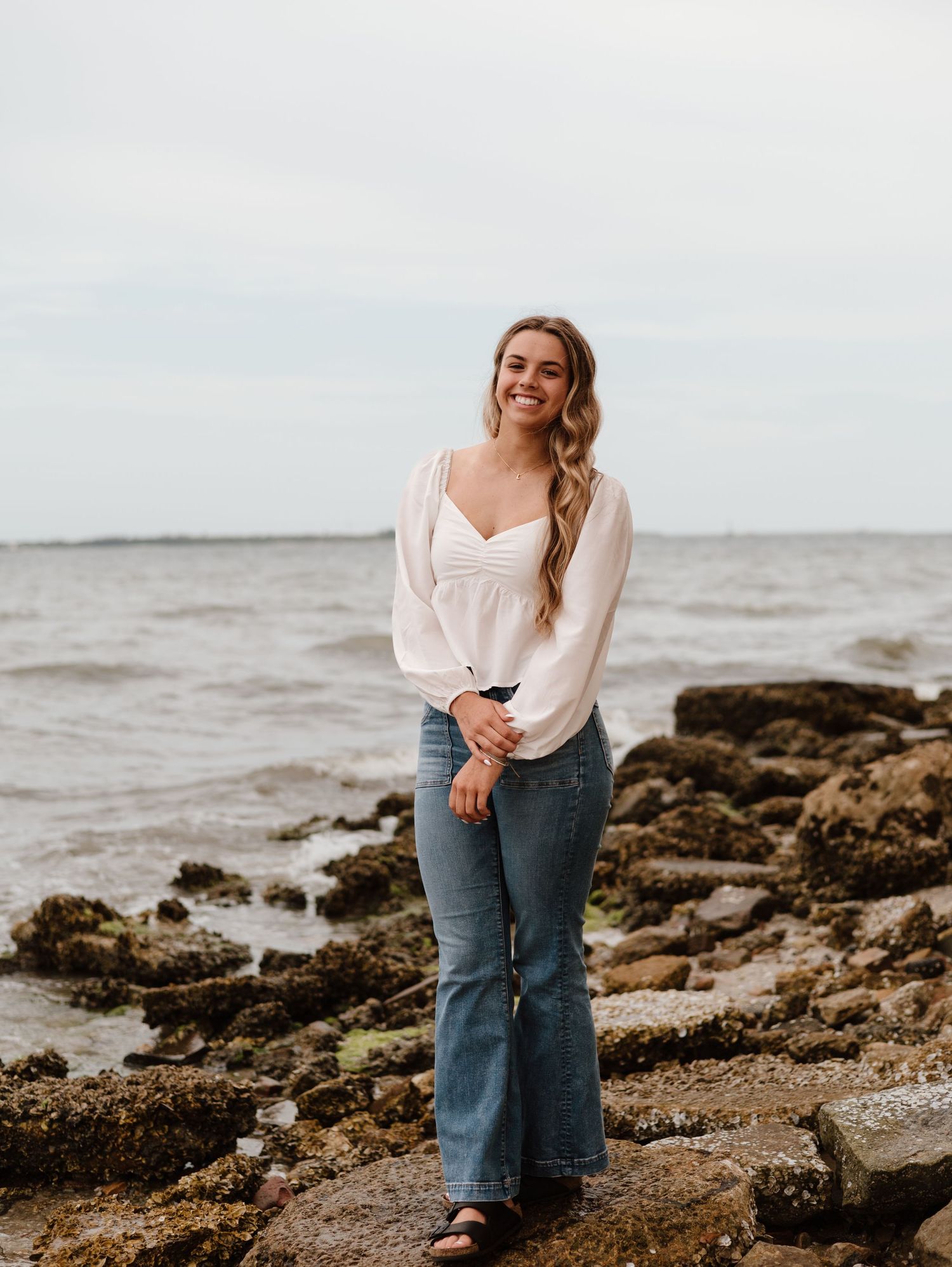 Someone wearing a white top and jeans poses casually on a rocky beach with the ocean in the background.