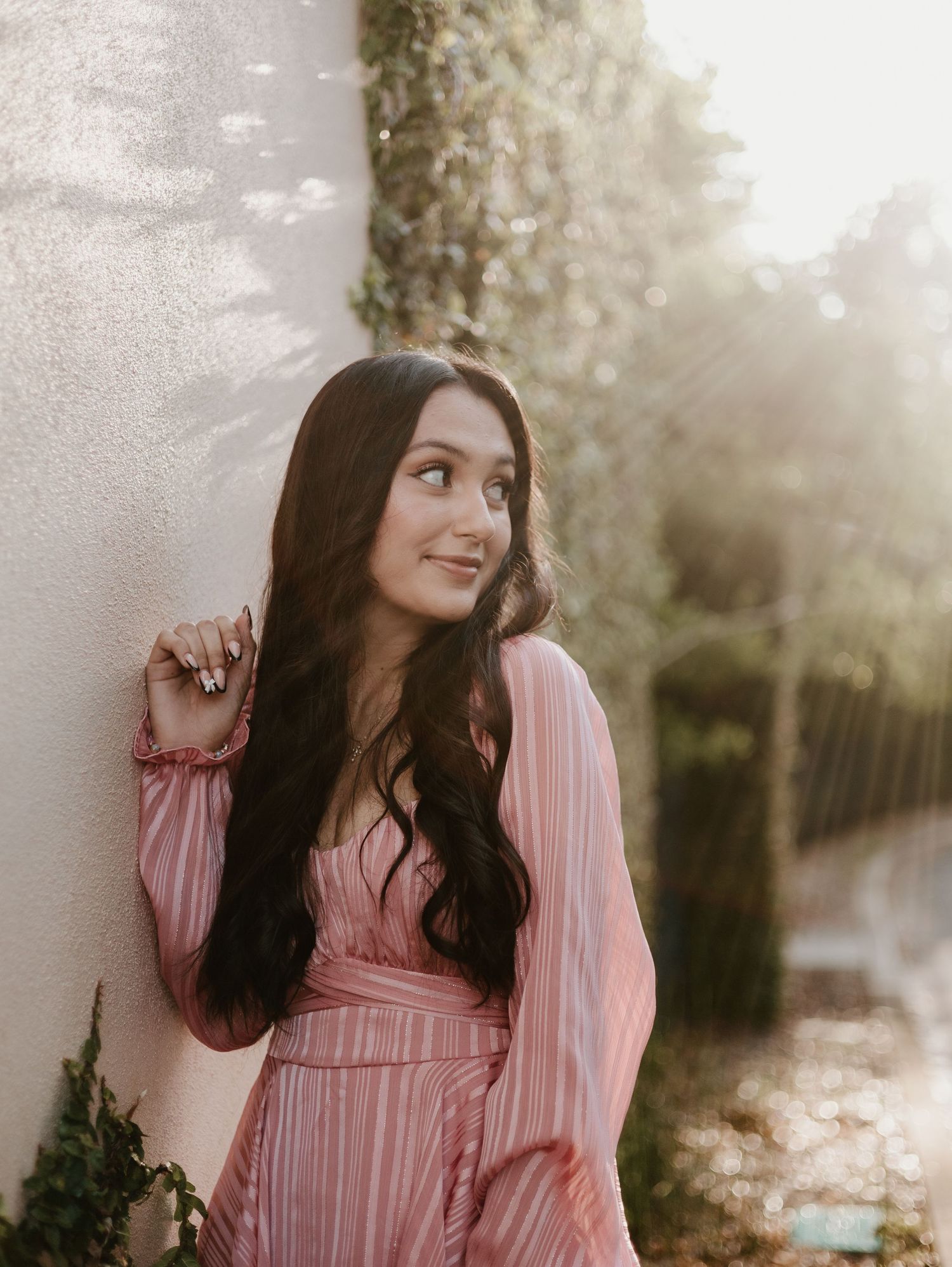 A woman in a pink pleated dress poses against a sunlit backdrop with dreamy bokeh effect.
