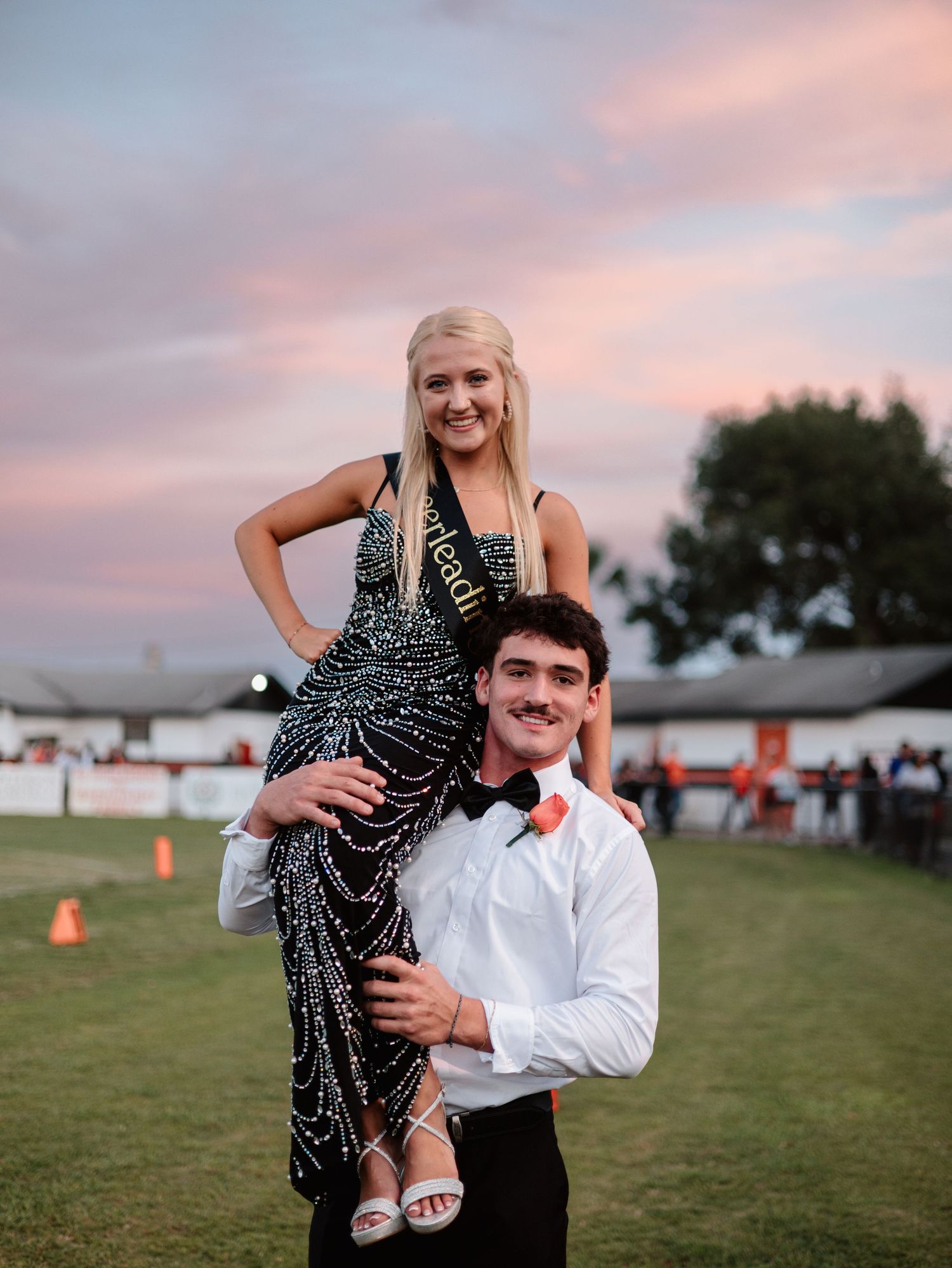 Couple shares playful moments on a grassy field during sunset with orange sky and trees in the background.