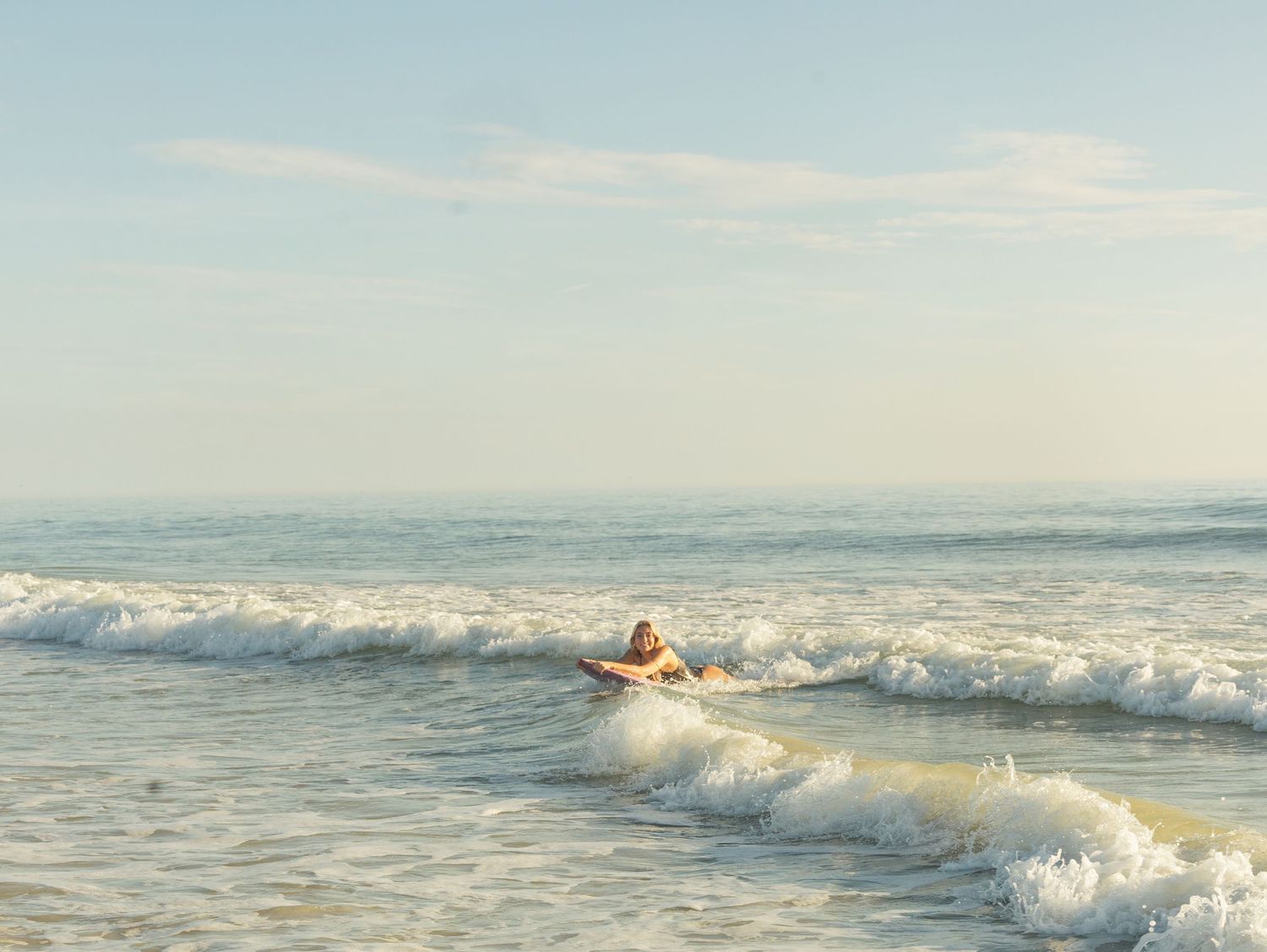 A surfer rides gentle ocean waves on a sunny day at a calm, picturesque beach.