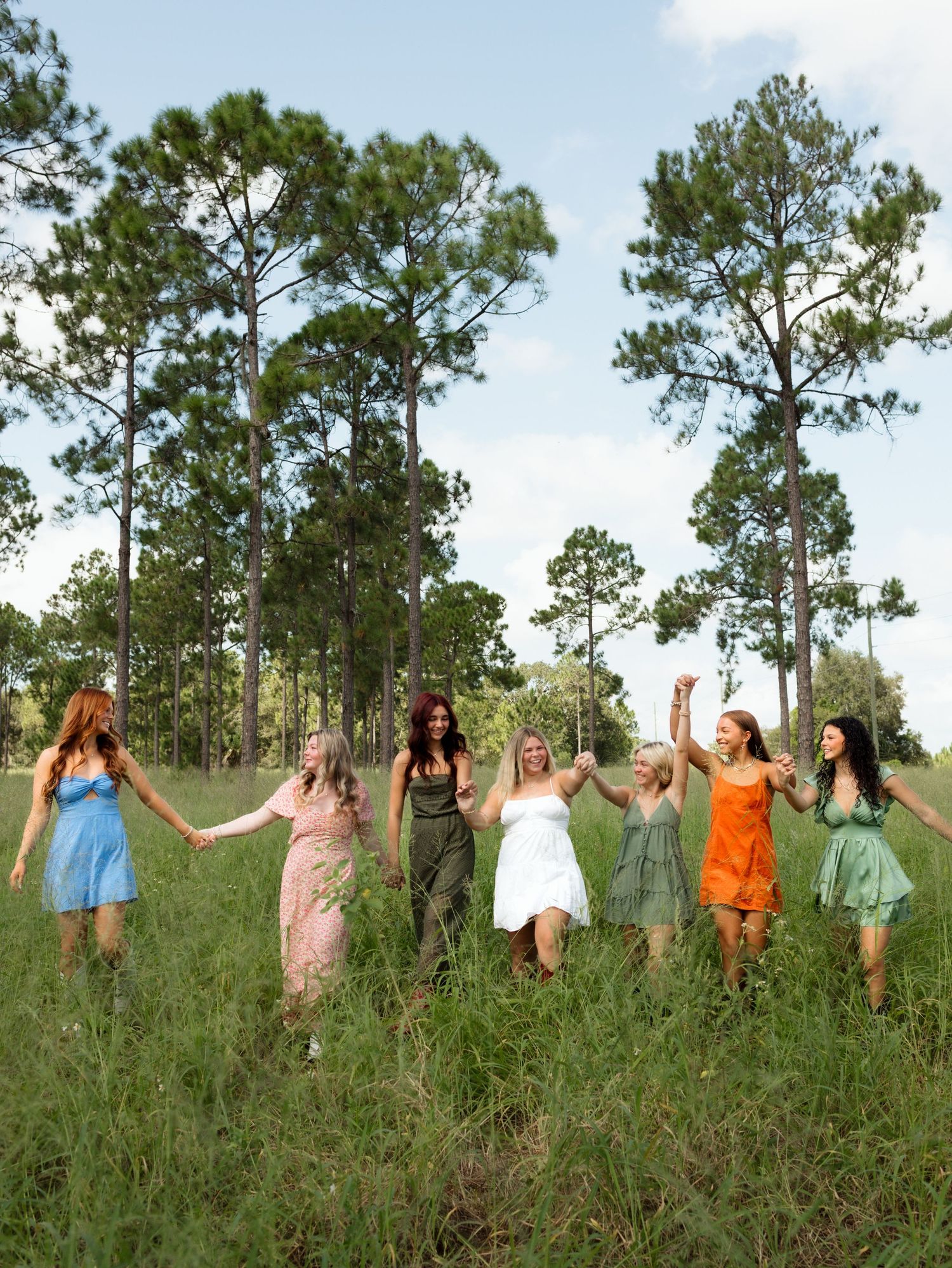 A group of people hold hands in a line while standing in a grassy field with tall pine trees in the background.