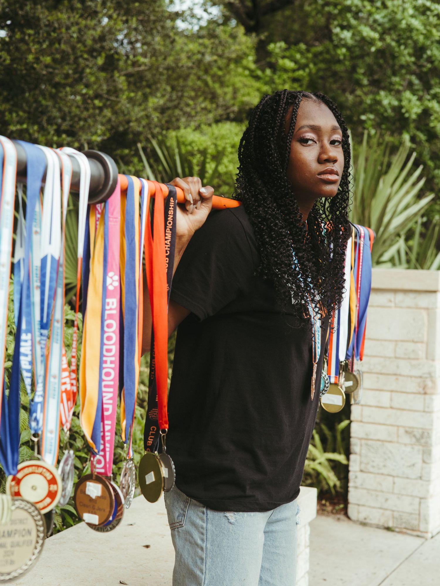 Person in black hoodie and jeans displays collection of colorful award medals and ribbons hanging on wall outdoors.