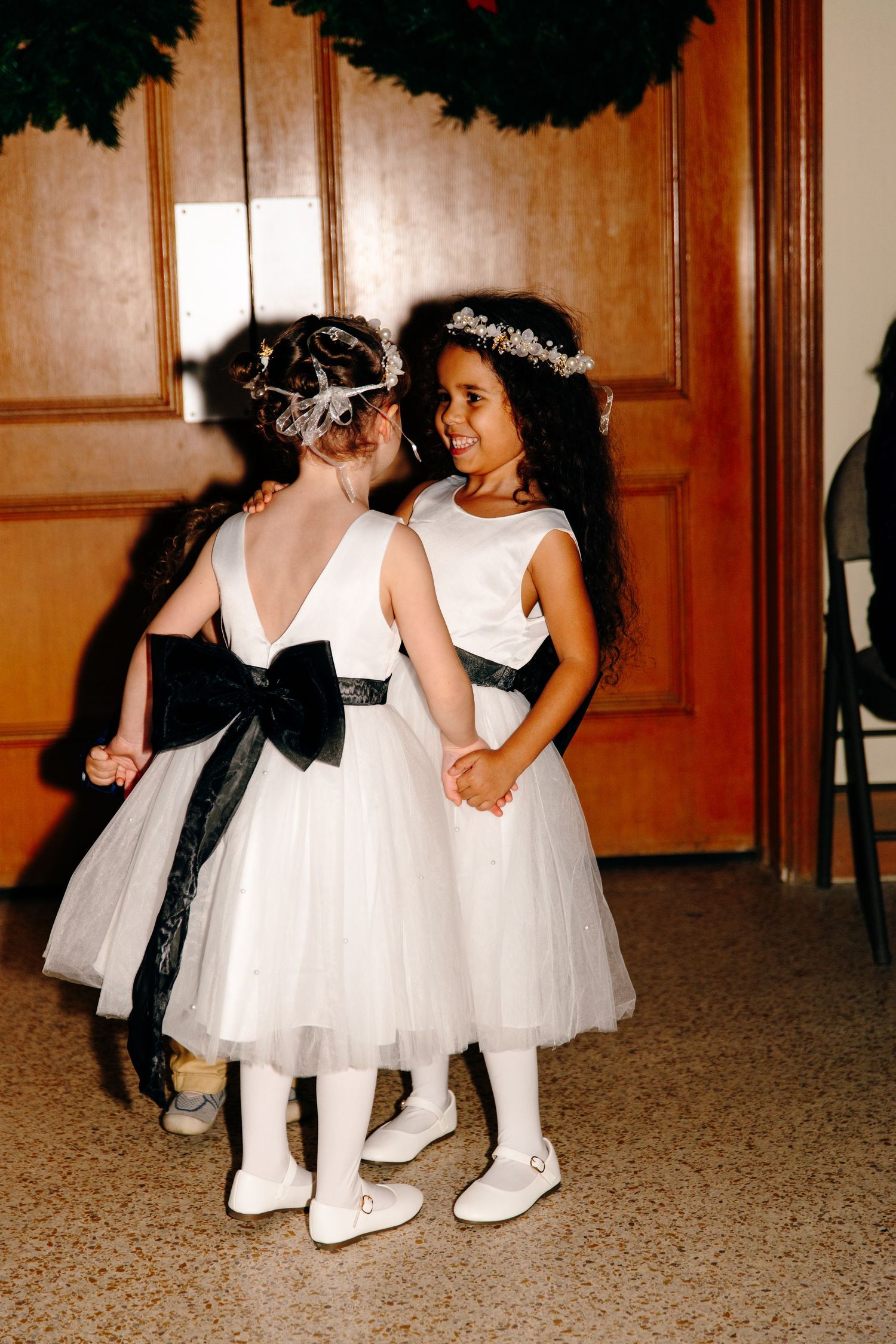Two flower girls in white dresses with black sashes and floral headpieces holding hands at a wedding.