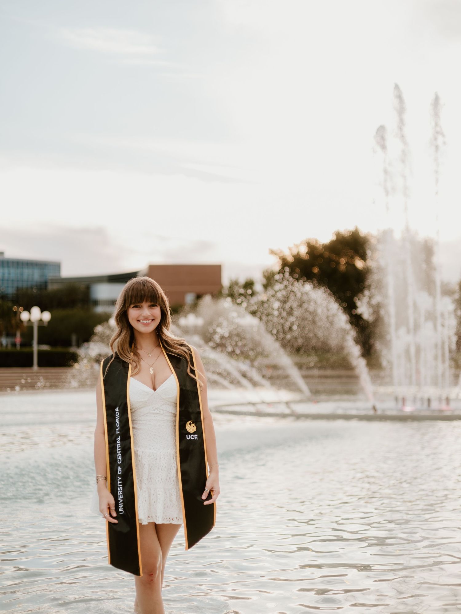 A smiling graduate wearing a UCF stole poses in a white dress near a fountain on a sunny day.