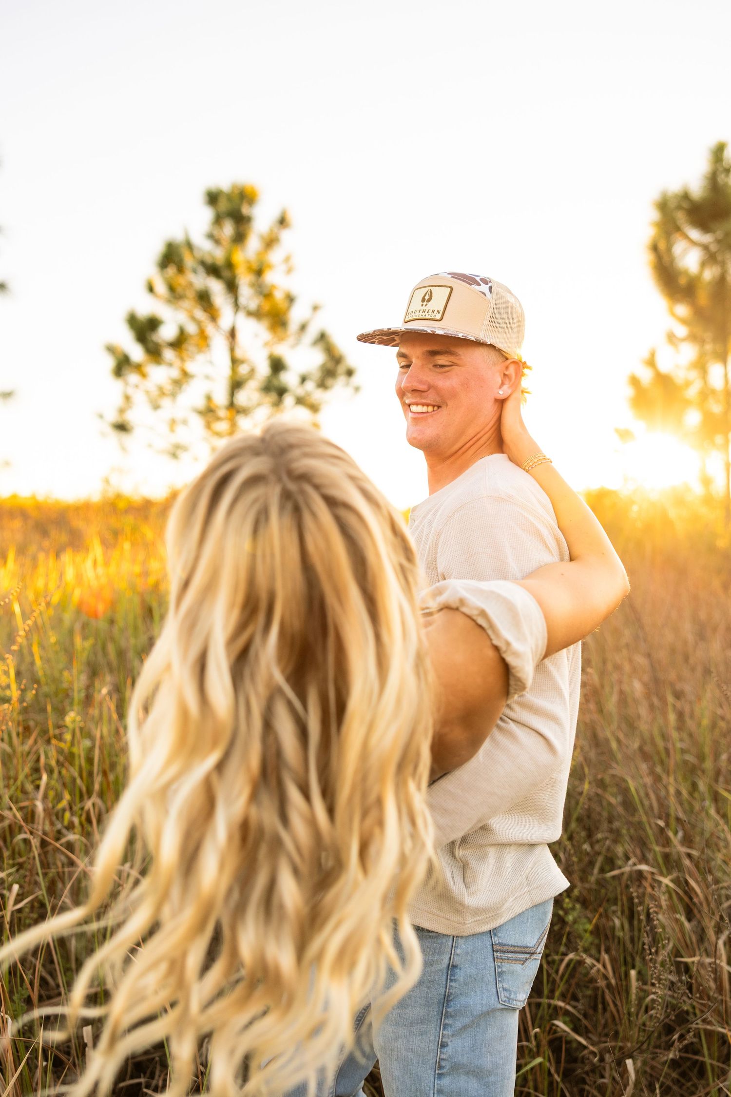 A smiling young man in a cap and jeans holds a blonde woman close in a sunlit golden field at sunset.