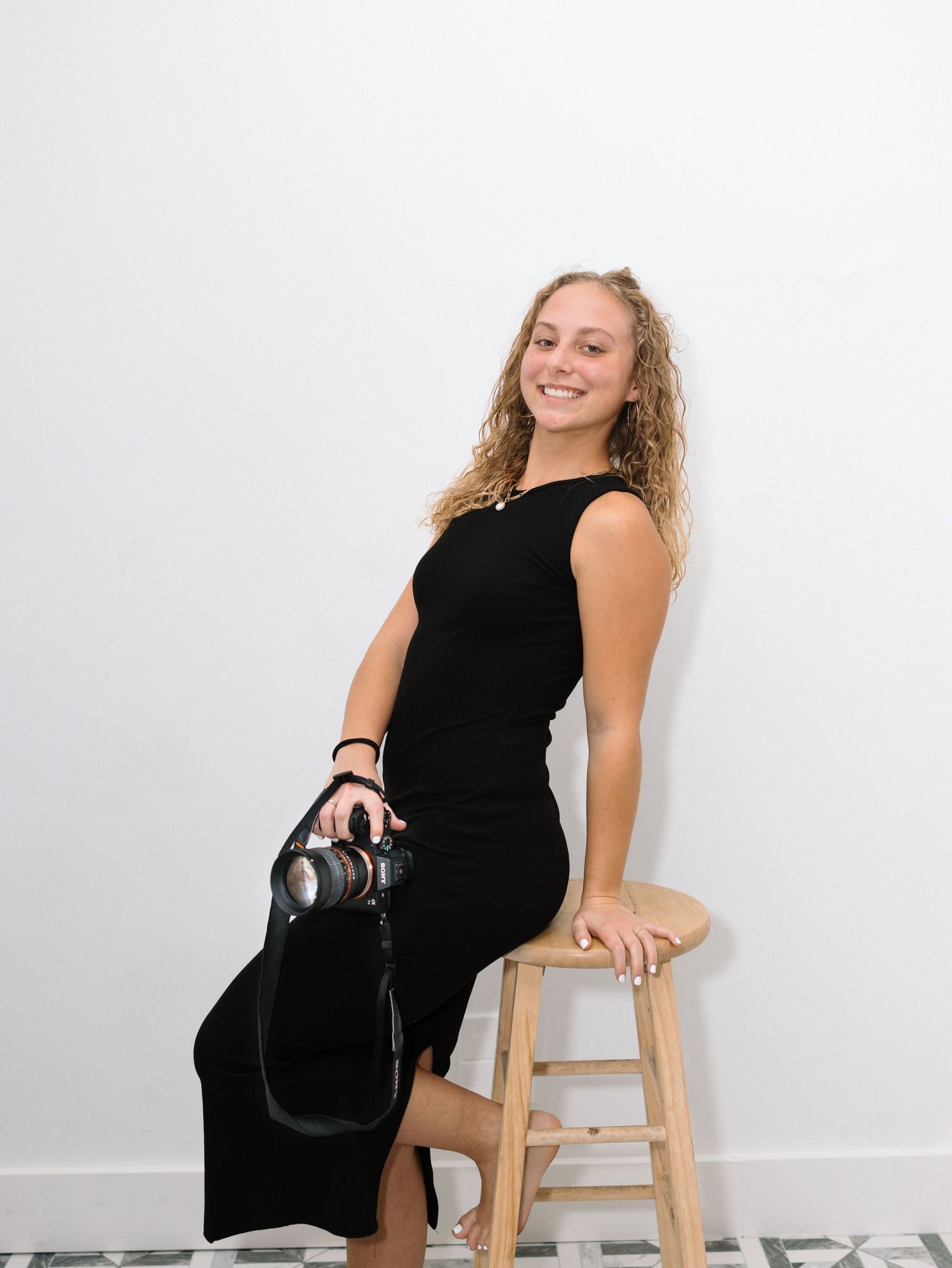 Young woman in black dress holding a camera, smiling while seated on a wooden stool against a white wall.