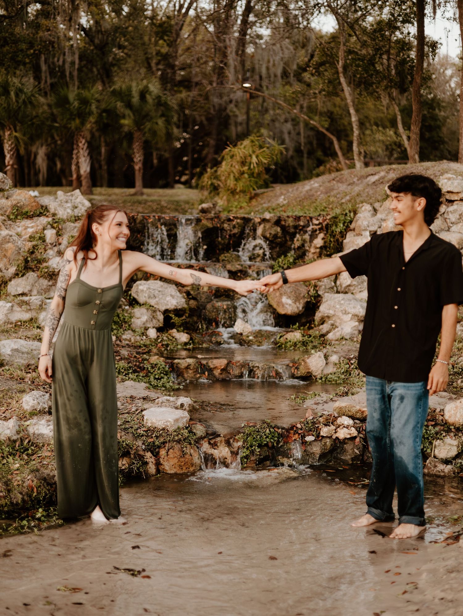 Two people hold hands in front of a cascading waterfall and rocky landscape at dusk.