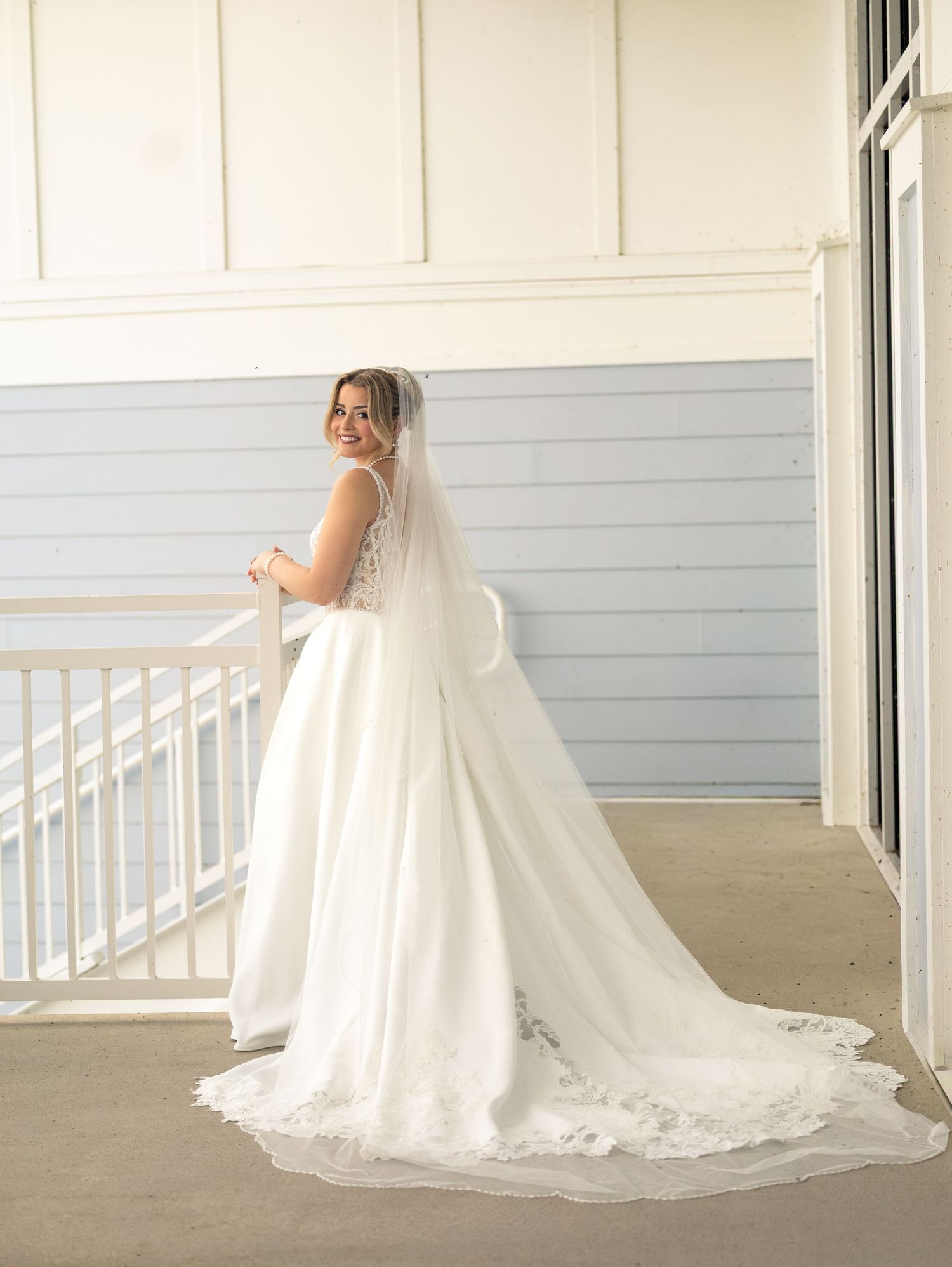 Bride in elegant white wedding gown with lace-trimmed cathedral veil standing on a balcony, looking back over her shoulder.