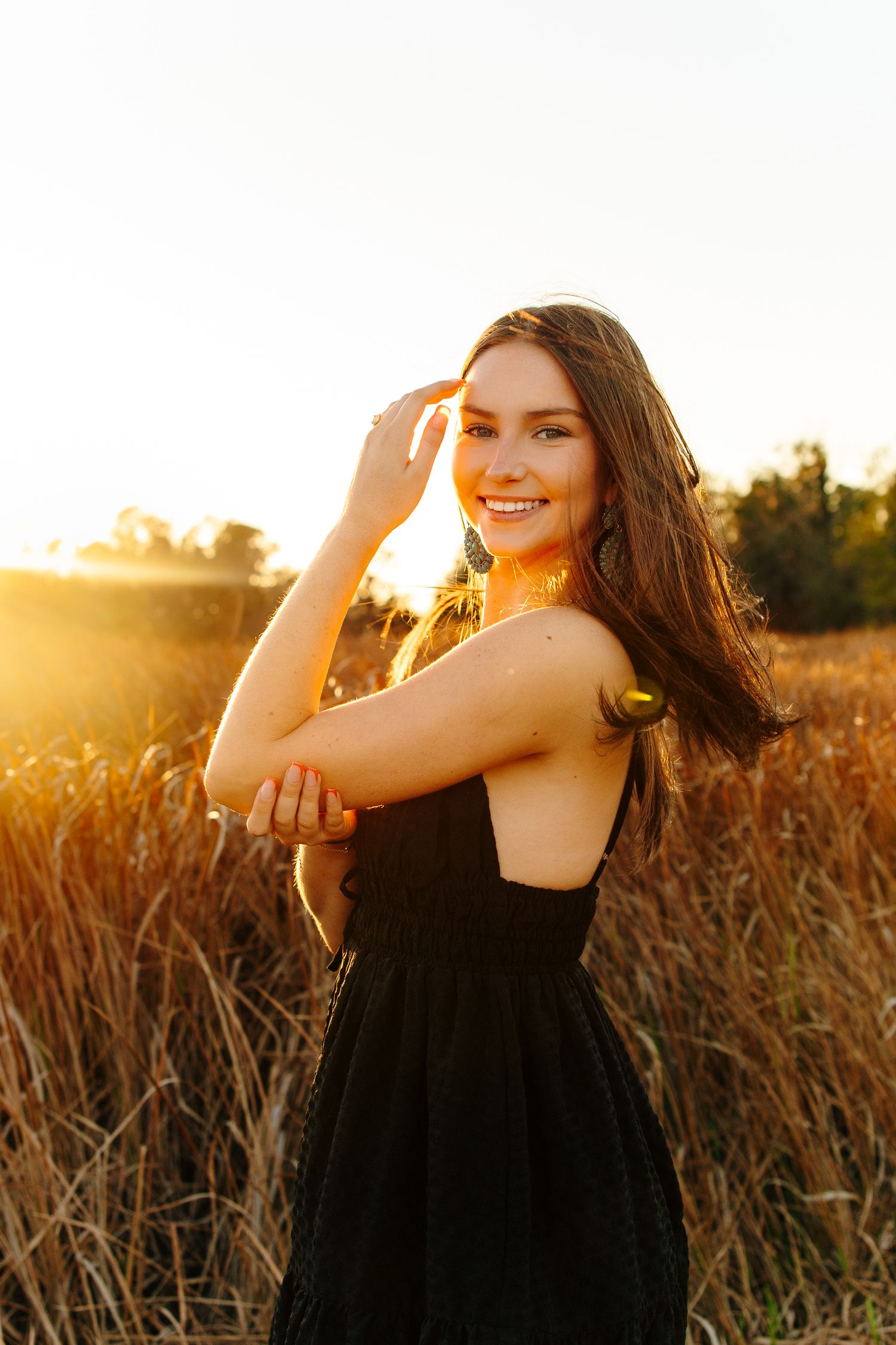 Young woman in black dress smiling in golden field at sunset, long brown hair, warm backlit glow.