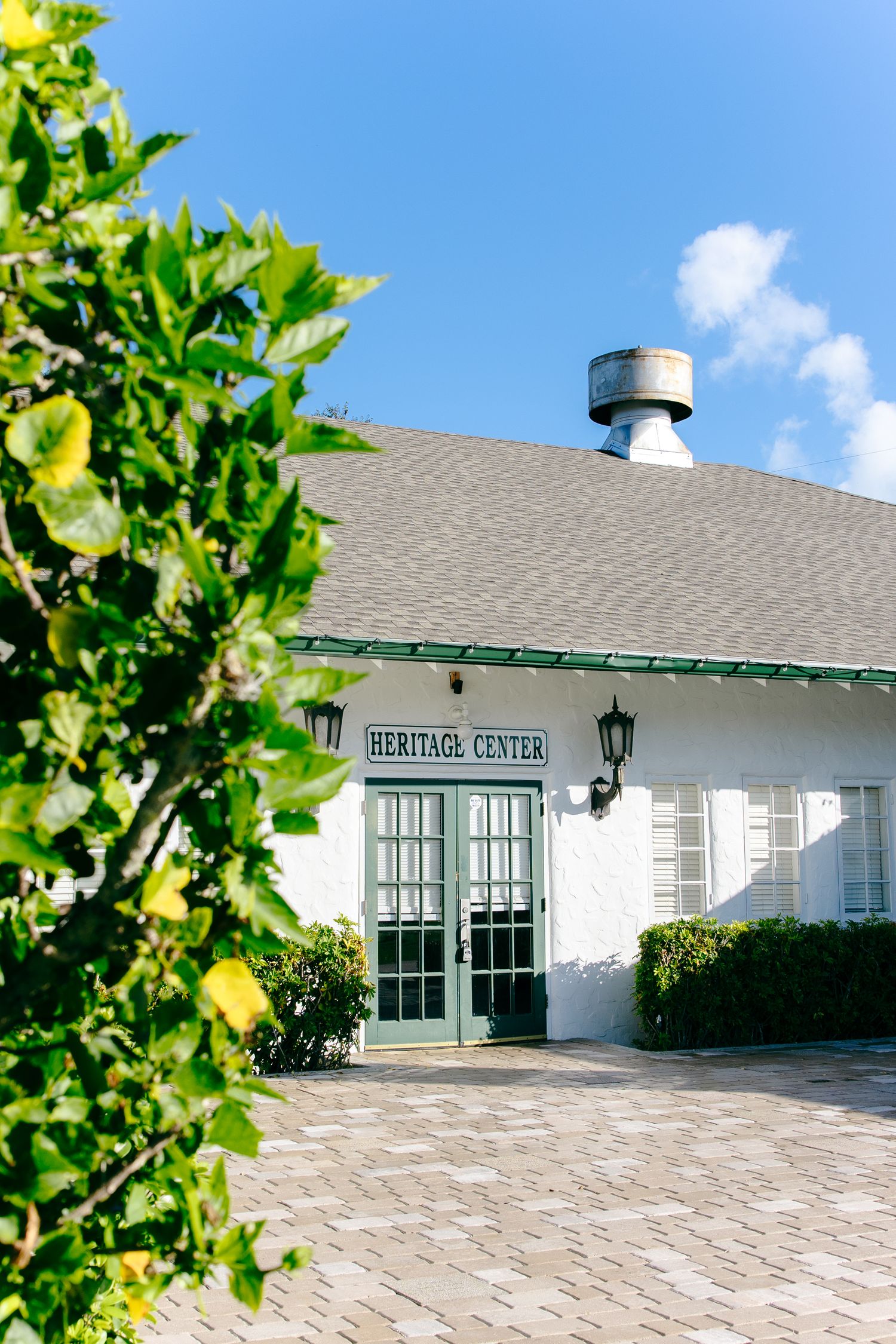 White Heritage Center building with green trim, brick patio, and lush greenery under a bright blue sky.