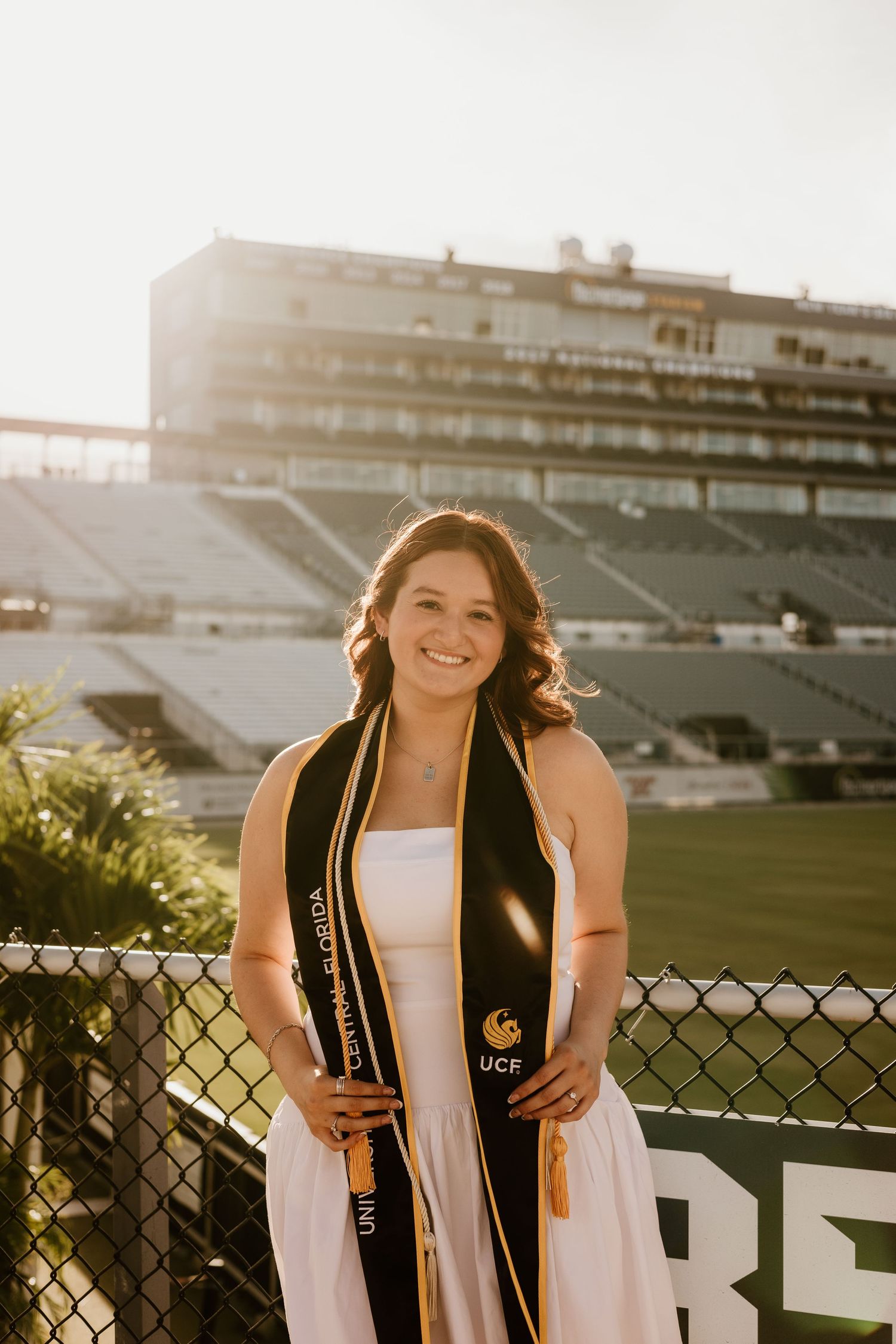 UCF graduate smiling in white dress with black and gold graduation stole at Bounce House stadium.