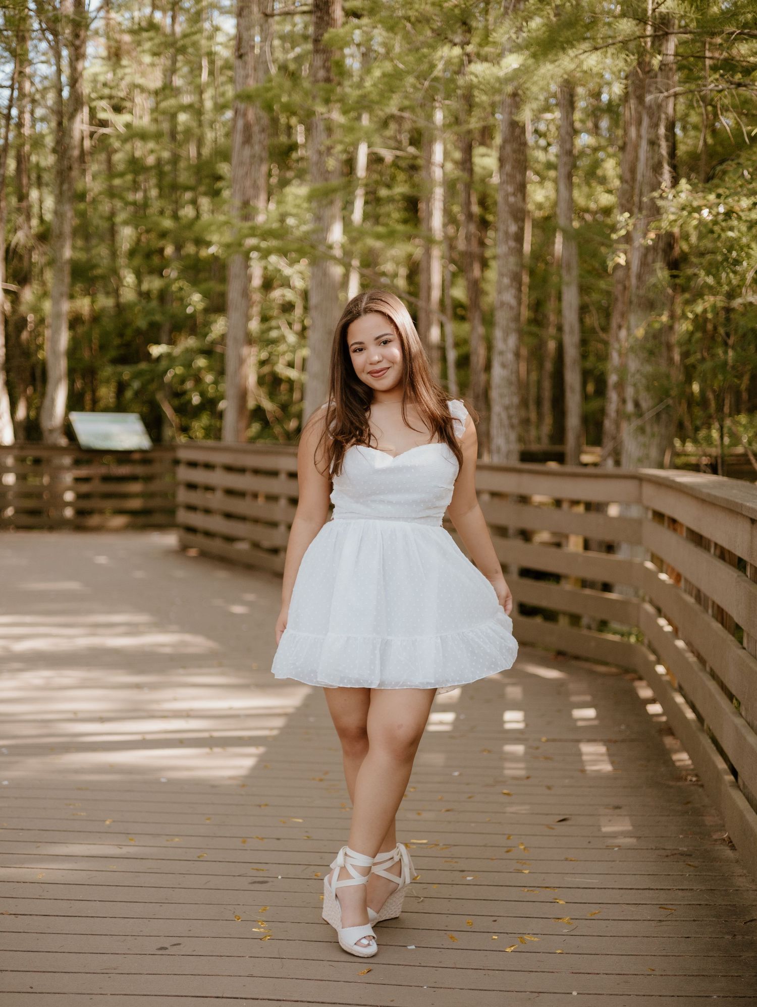 Young woman in white mini dress and heels posing on wooden boardwalk surrounded by lush green forest.