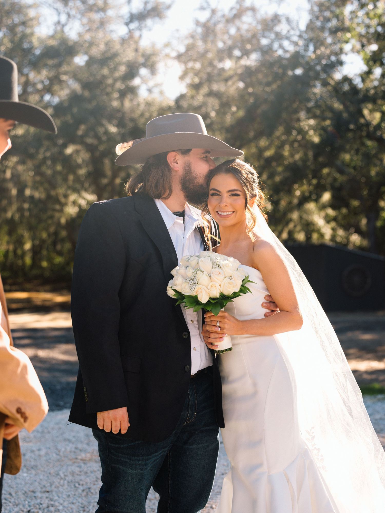Bride and groom in cowboy hats pose outdoors at rustic country wedding, bride holding white rose bouquet in elegant gown.