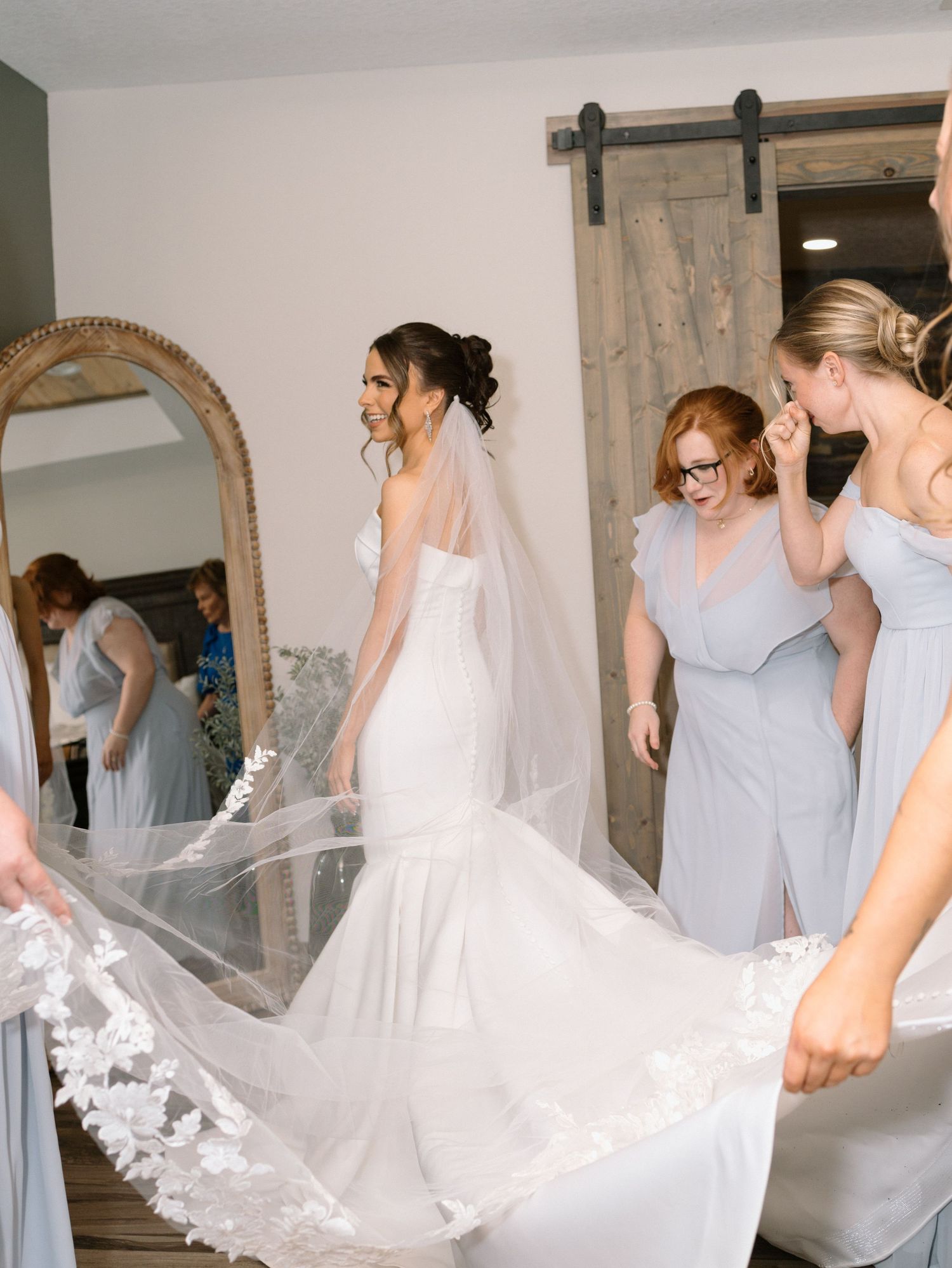 Bride in mermaid gown with lace veil surrounded by bridesmaids in blue dresses preparing for wedding day.